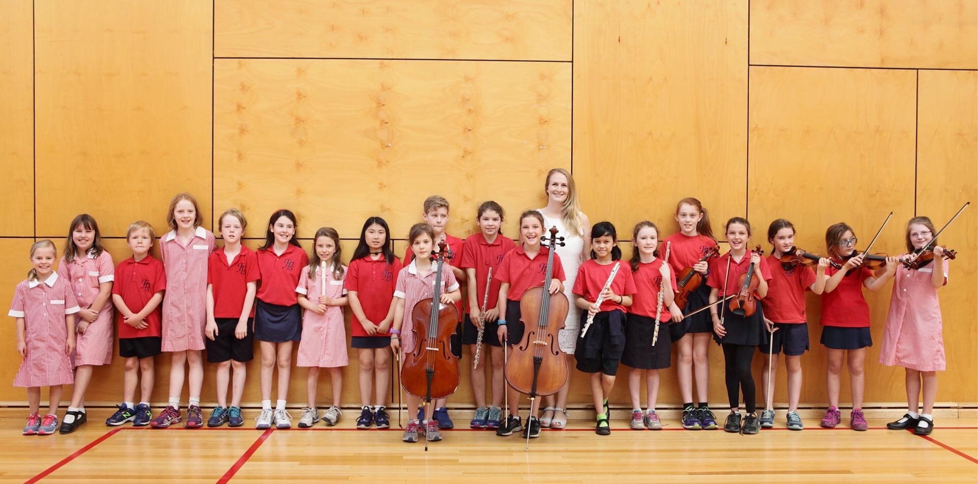 Group of young children and a woman, possibly an music teacher, standing in a line in a gymnasium with a wooden wall background. Some children are holding musical instruments, including violins, flutes, and cellos.