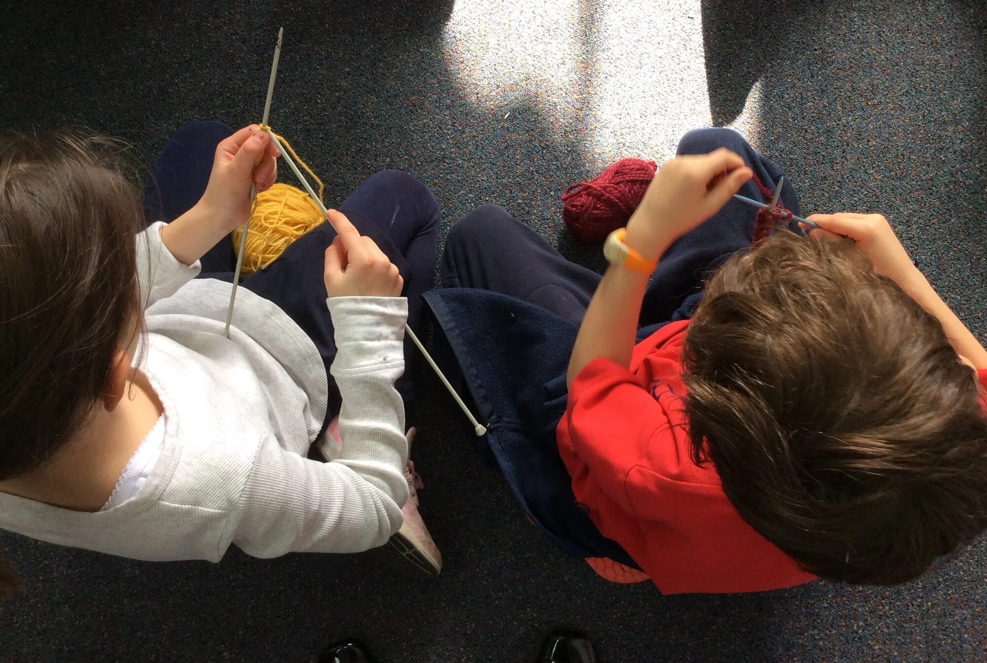 Two children sitting on a carpeted floor knitting with yarn.