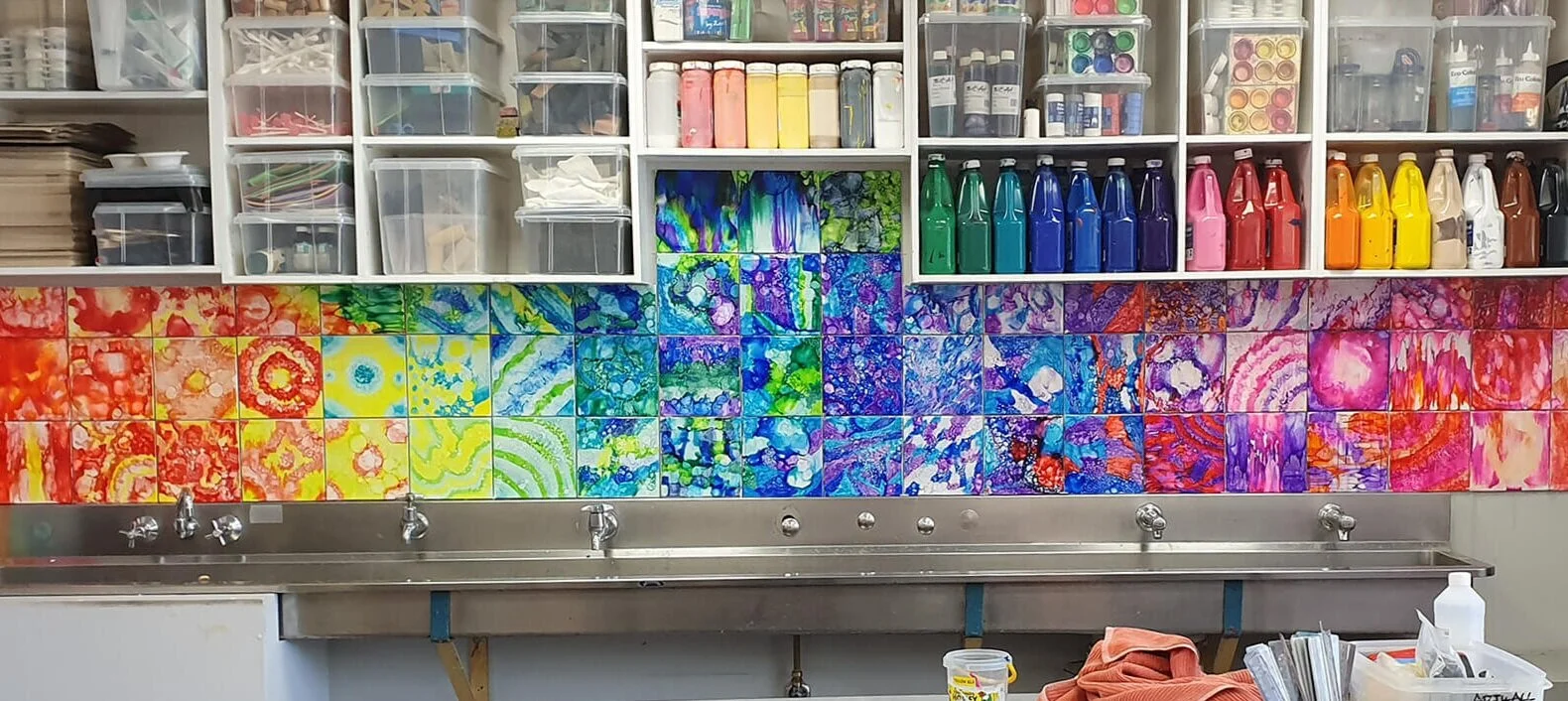 A colorful mural of abstract, vibrant paintings in various bright colors on a workshop wall behind a stainless steel sink with faucets, plastic storage bins, and art supplies.