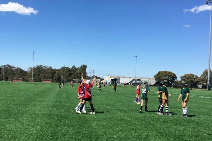 Kids playing on a soccer field, some wearing red and others in green uniforms, under a blue sky with a few clouds.