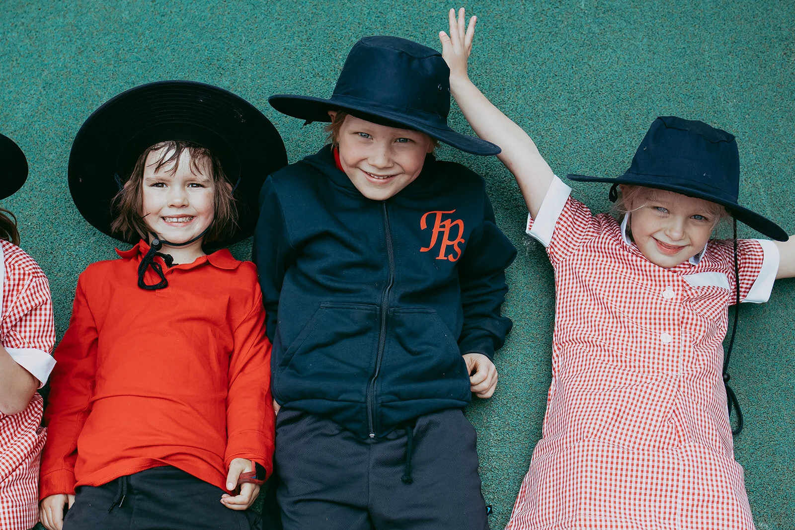 Three young children wearing hats and casual clothing, lying on a green textured surface and smiling at the camera.