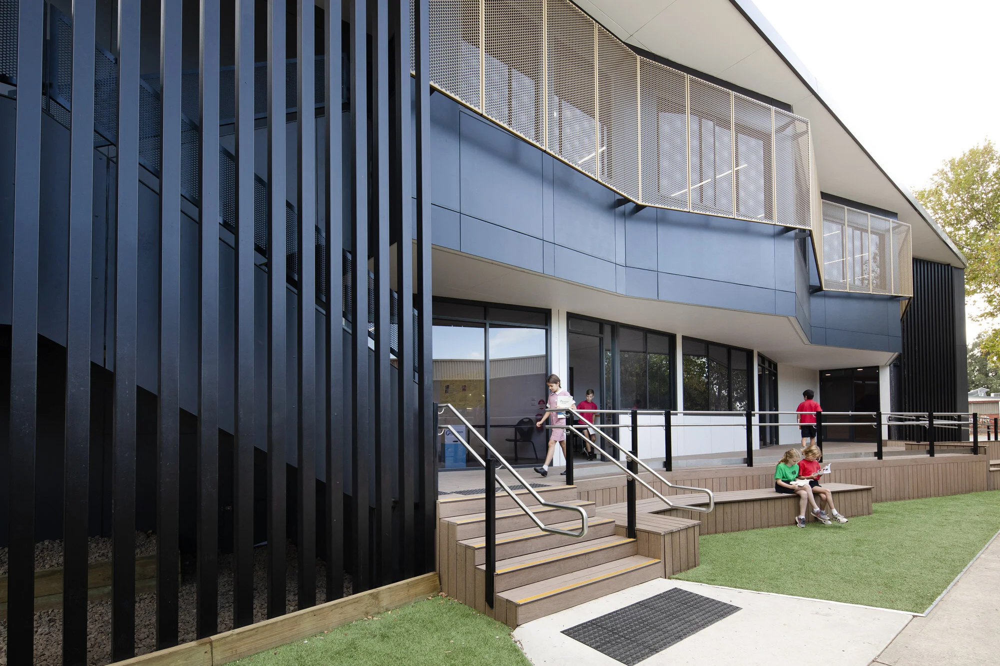 Modern school building with large glass windows, black vertical slats on the exterior, and a small outdoor seating area with children playing and sitting.