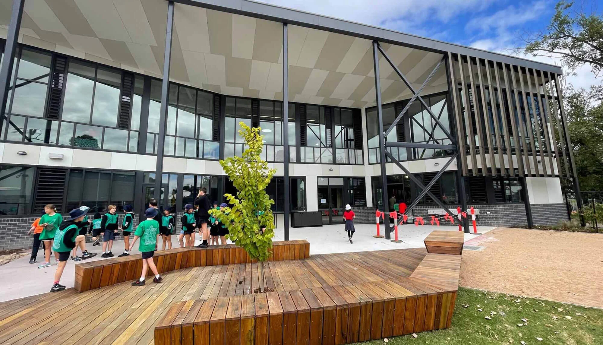 Students gather outside a modern school building with large glass windows and black framing, some wearing green uniforms and hats, in a courtyard with wooden benches and a small tree.