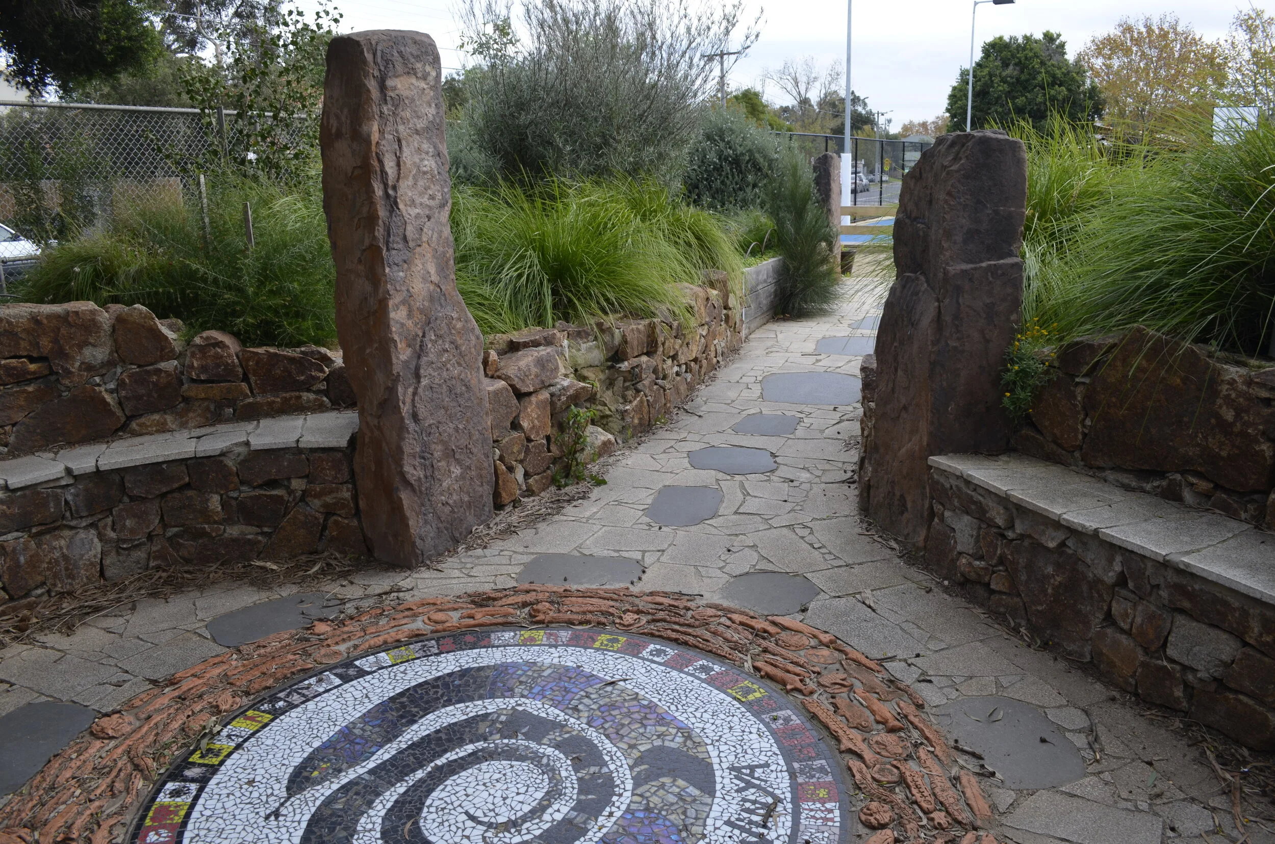 A stone pathway entrance with large upright rocks on either side, flanked by stone walls and lush green plants, leading to a fenced outdoor area.
