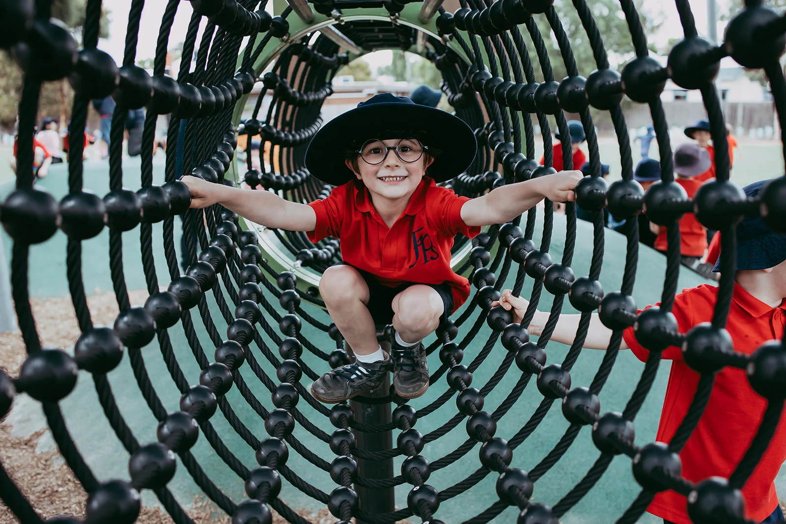 Child in red shirt and black hat crawling through a large black rope tunnel at a playground, smiling at the camera.