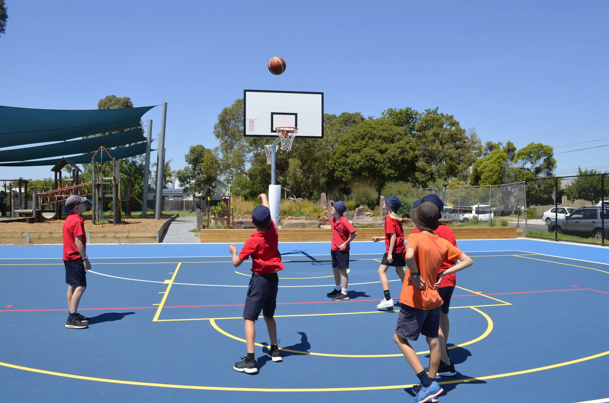 Children playing basketball on an outdoor court, with blue surface and yellow lines, under a clear blue sky.