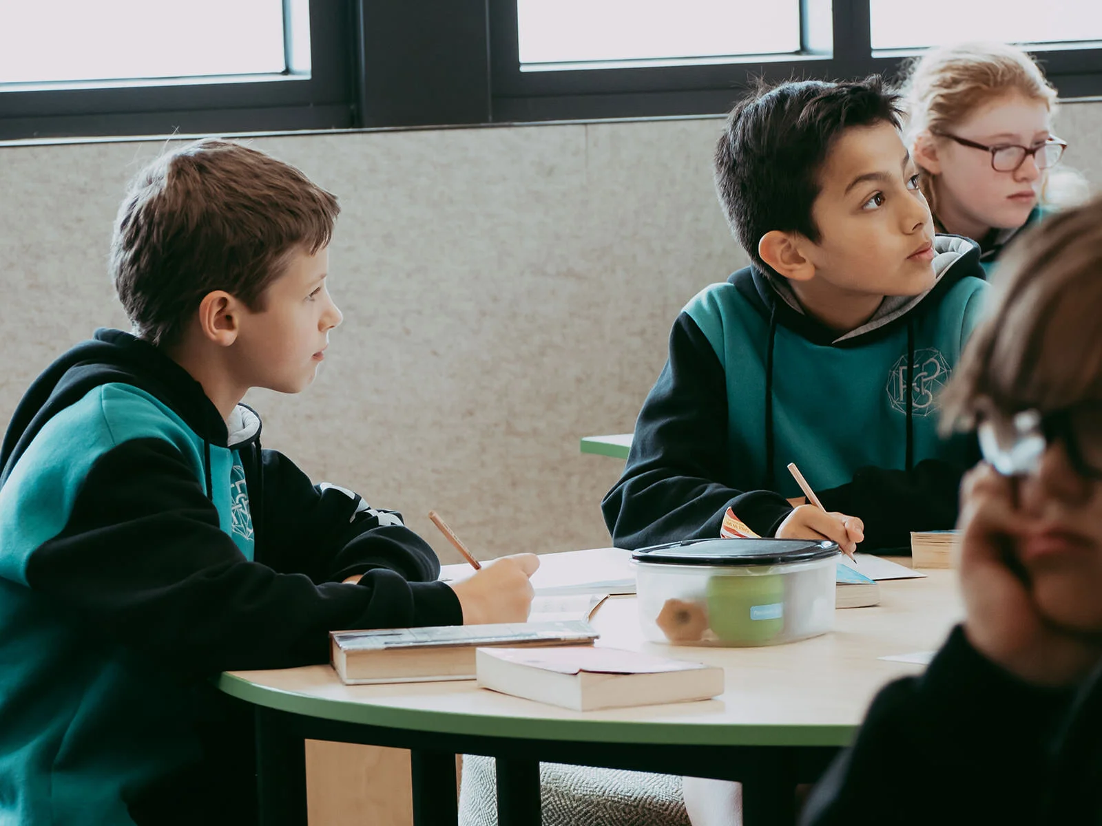 Four children in a classroom sitting at a round table, attentive and taking notes, with books and a container on the table, near large windows.