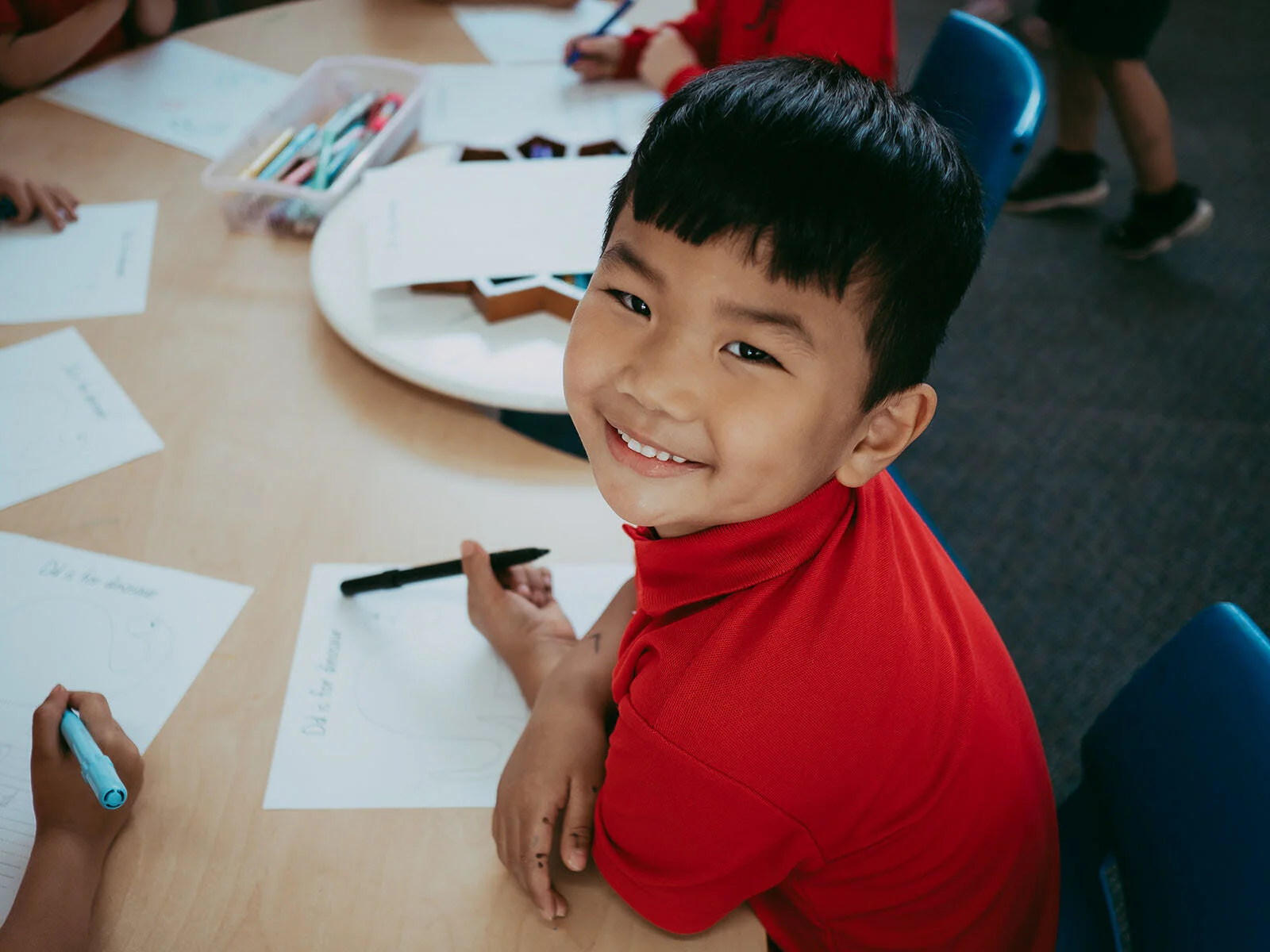 Smiling young boy in a red shirt sitting at a desk with papers and markers, working on an activity in a classroom.