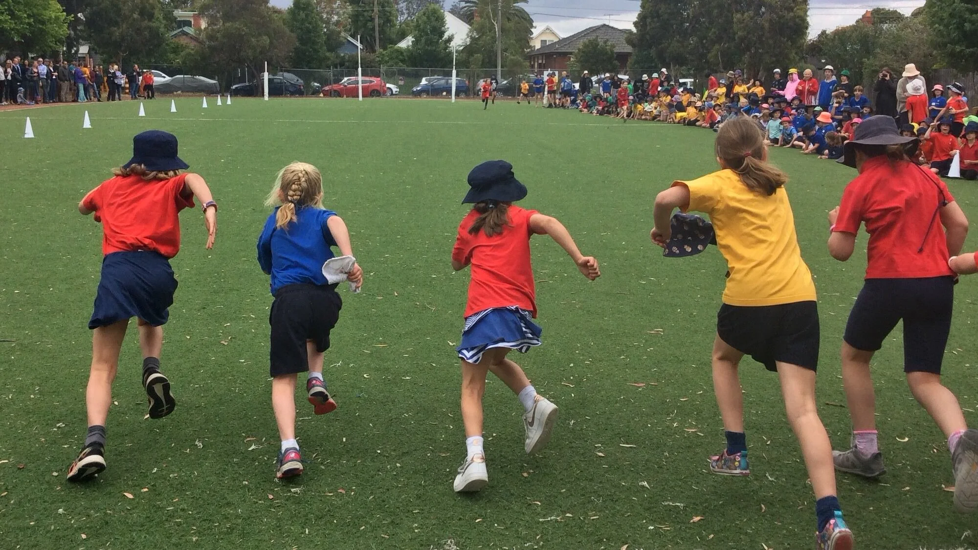 Children racing on a grassy field at a school sports day, with an audience of spectators seated and standing along the edge of the field.