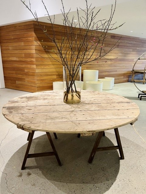 An large, round wooden table sits in the James Perse showroom.  A vase with oversized branches sits on top of the table.