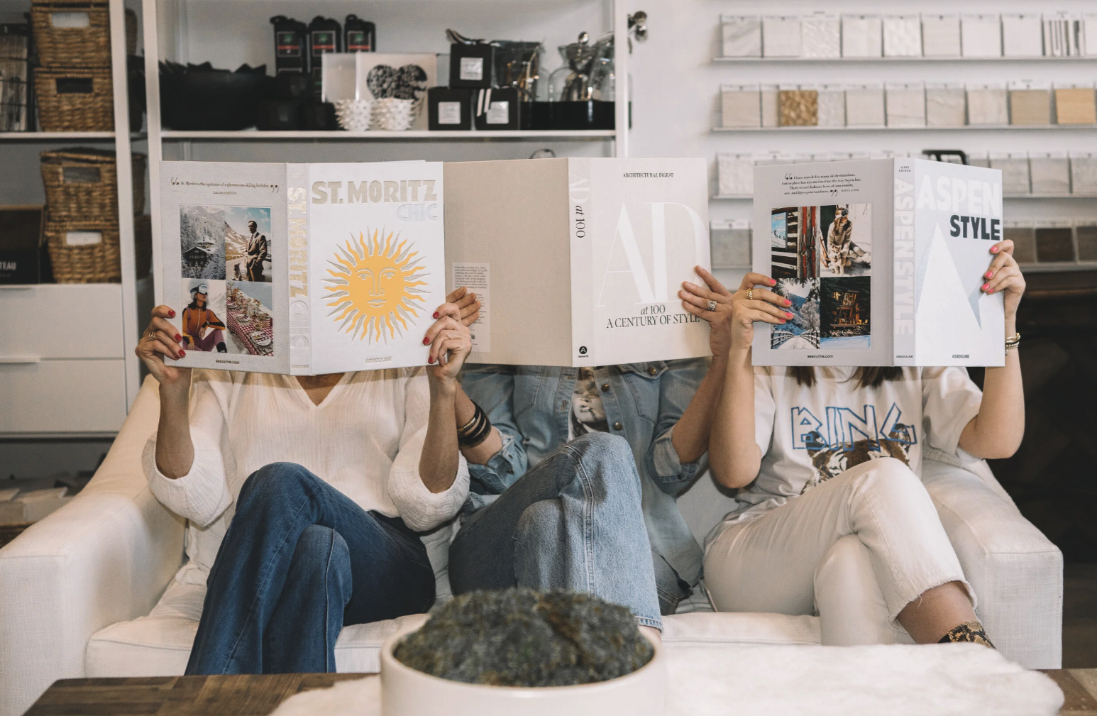 Three women sitting on a white couch, reading interior design books covering their faces, in a cozy decor store with shelves of samples and books in the background.