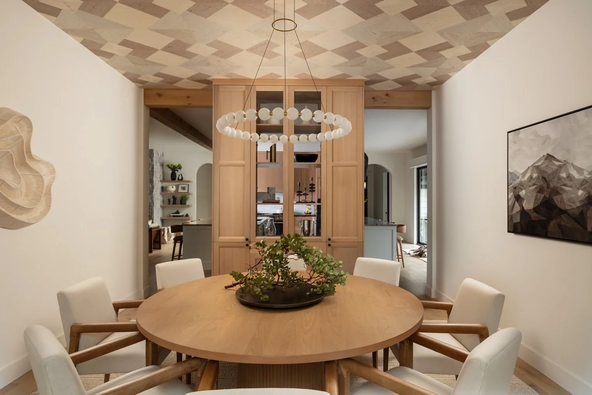 A dining room with a round wooden table and six white cushioned chairs. A modern pendant chandelier hangs above the table. There is a wooden cabinet with glass doors behind the table, and a decorative plant on the table. The walls are white, and the ceiling has a geometric pattern.