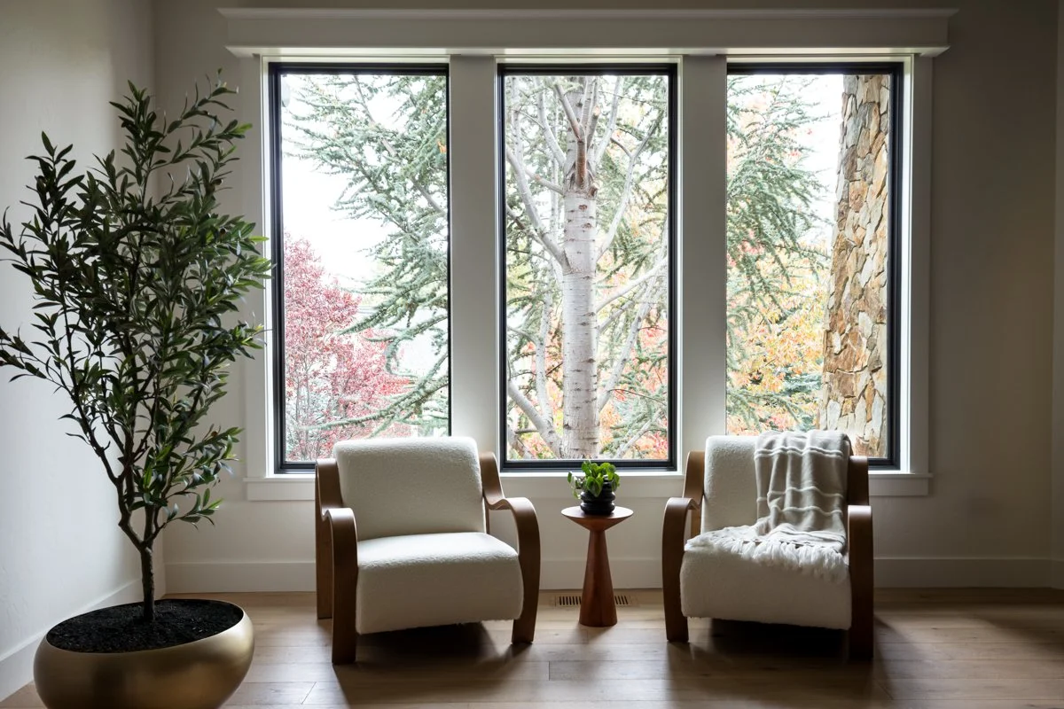 Luxury interior design by Design Bar Bend in Bend, Oregon for the ParkHaus project featuring a bright window seating nook with paired white bouclé armchairs, wood frame detailing, wood side table, indoor tree in gold planter, and floor-to-ceiling win