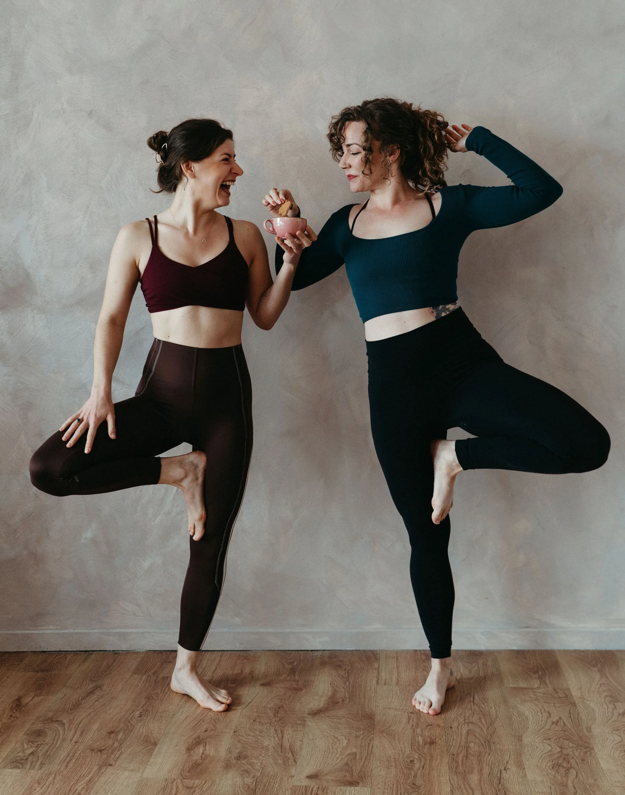 Two women practicing yoga together indoors, one holding a pink mug with cookies, both balancing on one leg in tree pose, smiling and enjoying each other's company.
