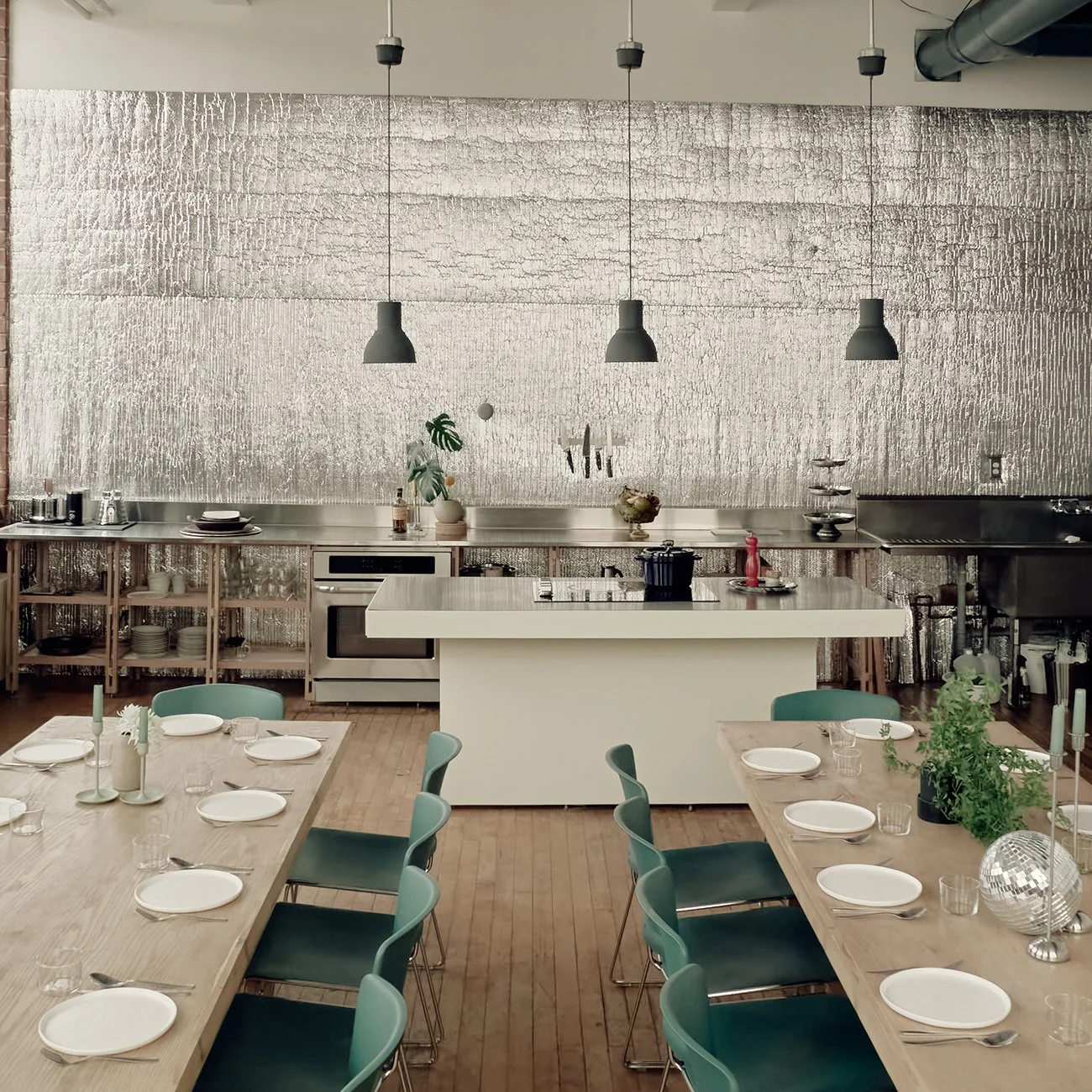 A modern kitchen and dining area with a metallic textured wall, wooden tables set with white plates and glassware, green chairs, and pendant lights hanging from the ceiling.
