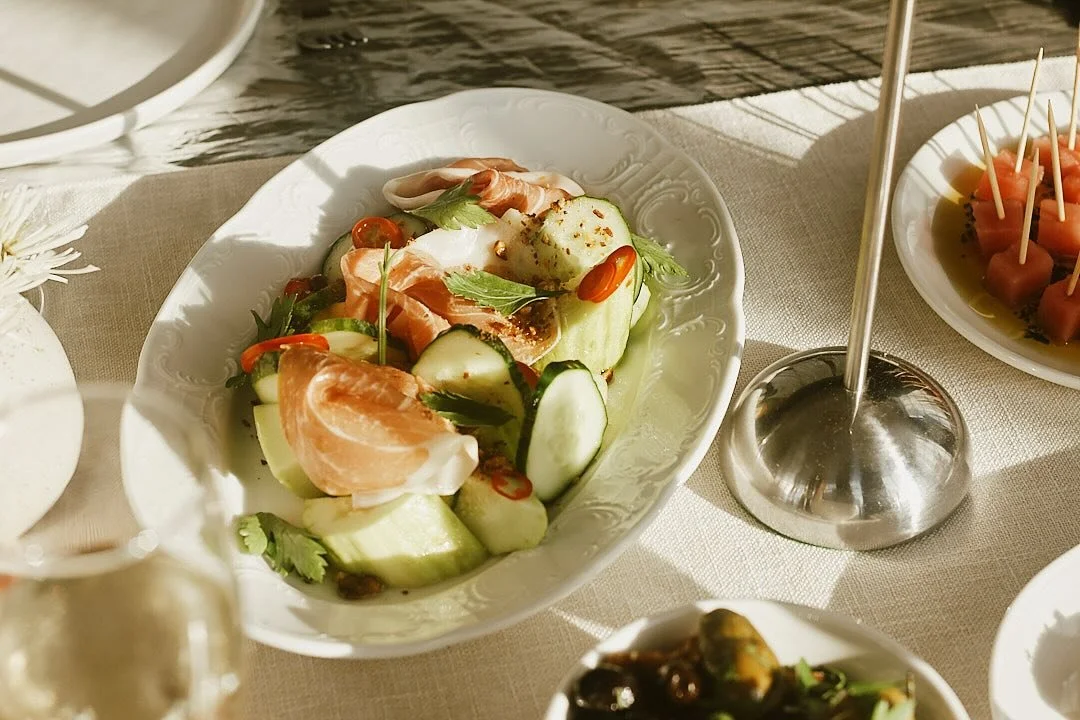 Fresh vegetable salad with cucumber, cherry tomatoes, red chili slices, and sliced cured meat on a white ornate dish, placed on a table with other appetizers.