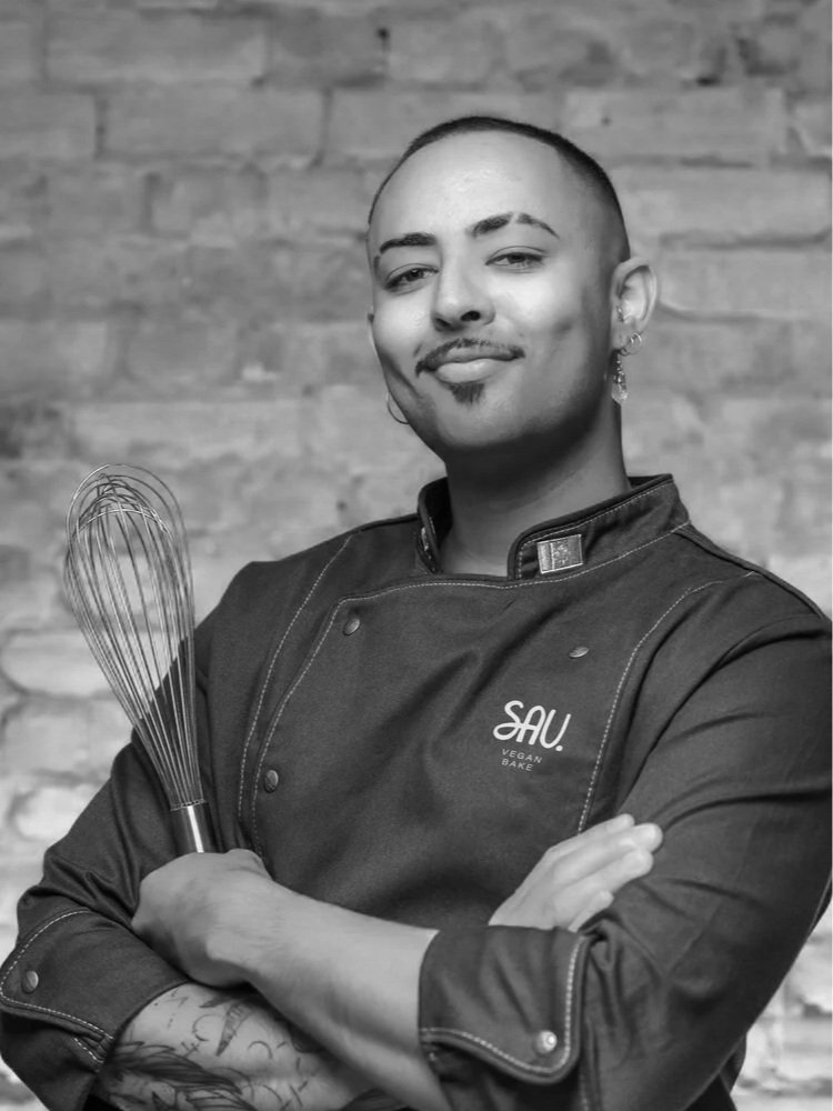 Smiling chef holding a whisk, wearing a dark jacket with 'SAU Vegan Bake' logo, against a brick wall backdrop.