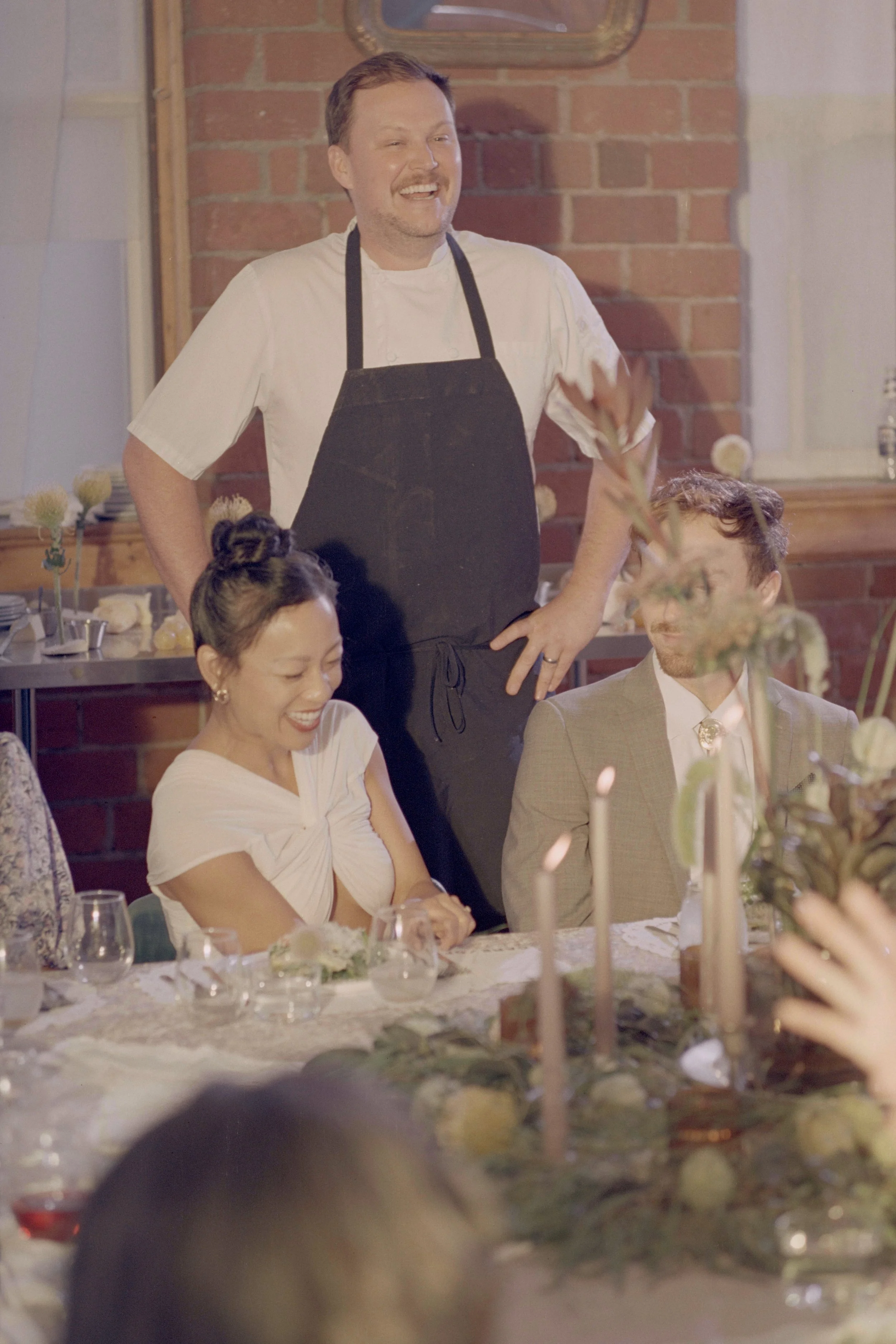 A man wearing a white shirt and black apron stands and smiles at a gathered group during a celebration. Seated are a woman with hair in a bun, wearing a white dress, and a man in a beige suit, at a table decorated with candles and flowers, in a room 