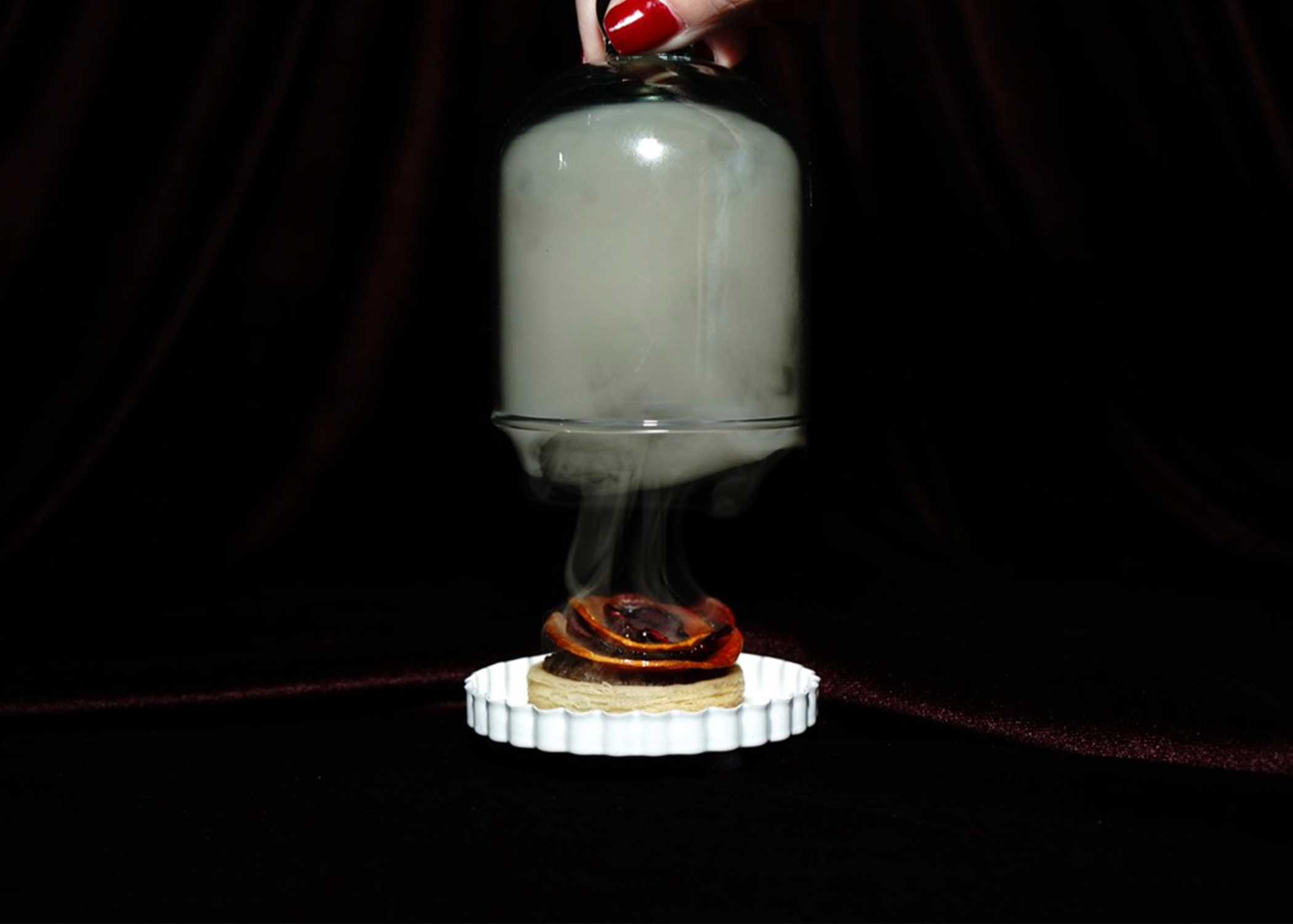 A glass container filled with a white, smoky substance is suspended upside down above a small dish. The dish contains a piece of bread topped with grilled red bell pepper and a brown item. Smoke or vapor is emanating from the dish, adding a dramatic effect, against a dark background with red fabric.
