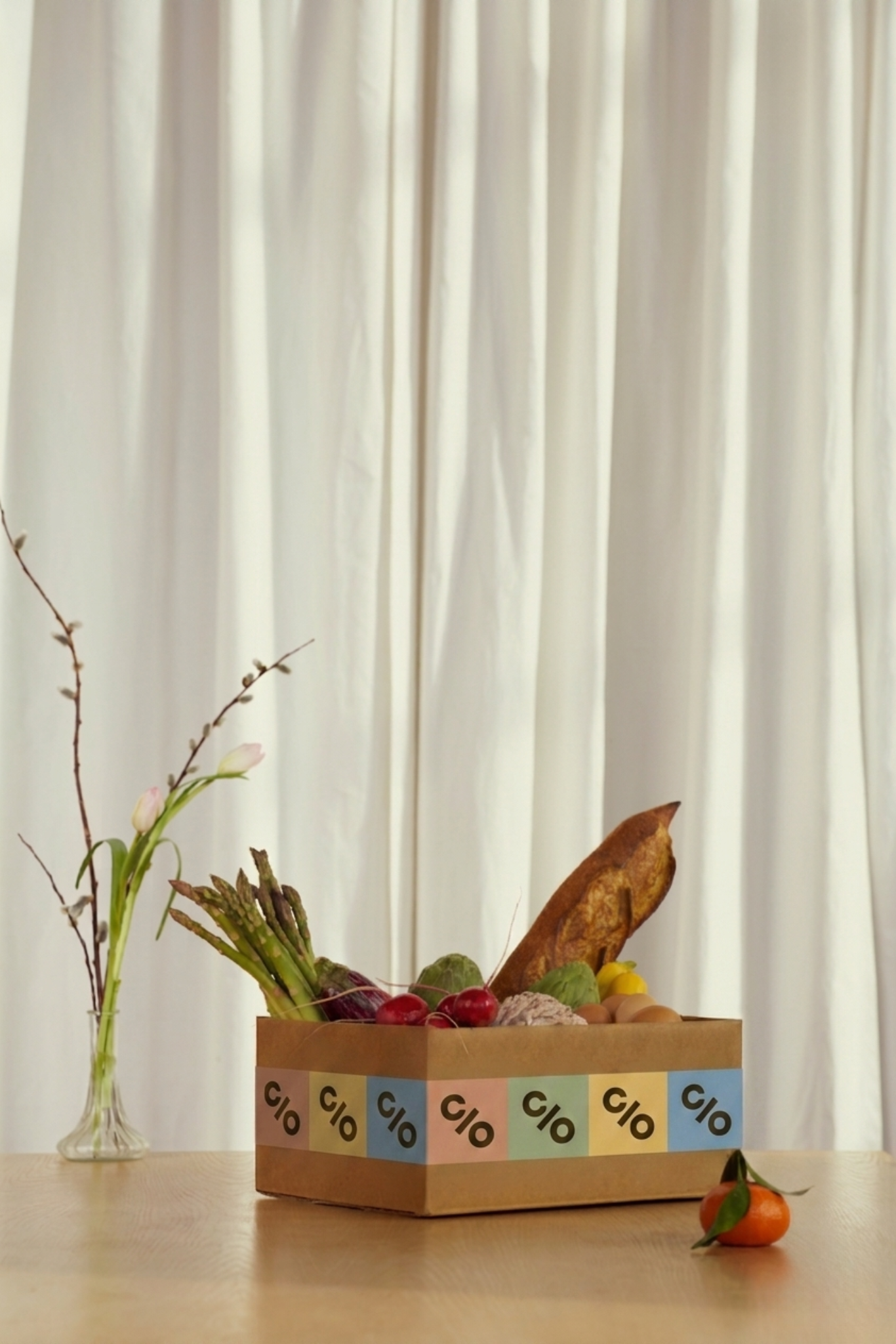 A cardboard box filled with various vegetables and fruits placed on a wooden table, with a clear vase holding branches and a flower to the left, and a tangerine with green leaves on the table to the right. The background features a cream-colored curtain.