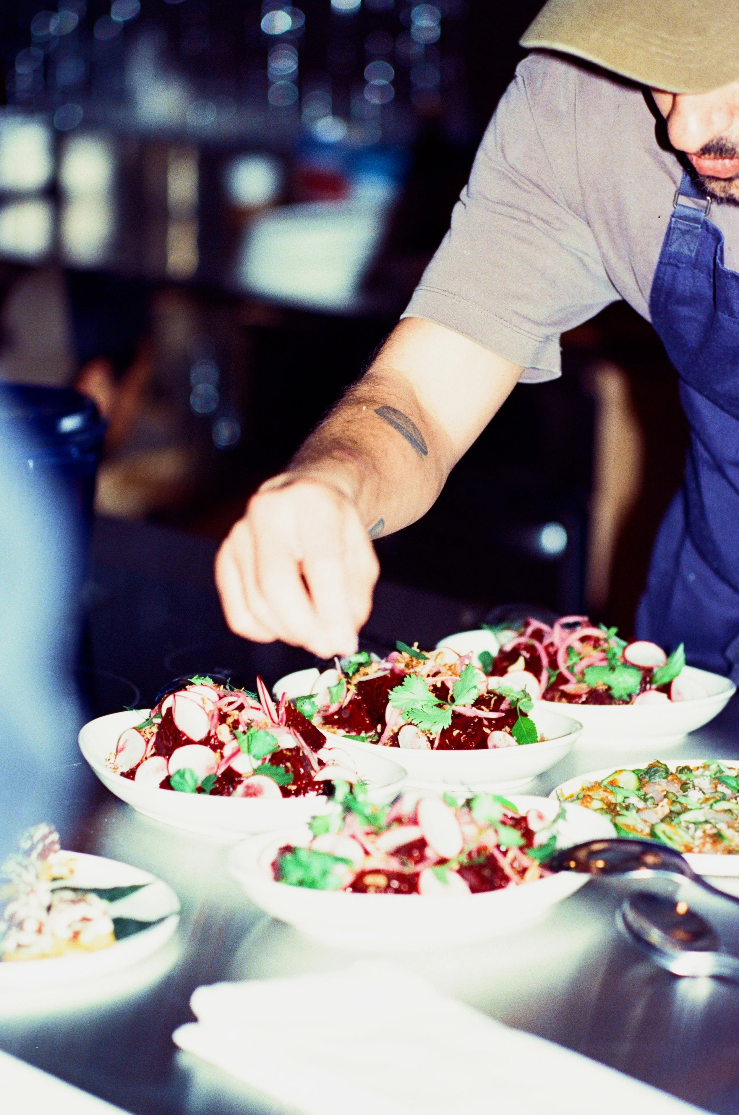 A chef preparing salads with radishes, beets, herbs, and other vegetables in white bowls on a kitchen counter.