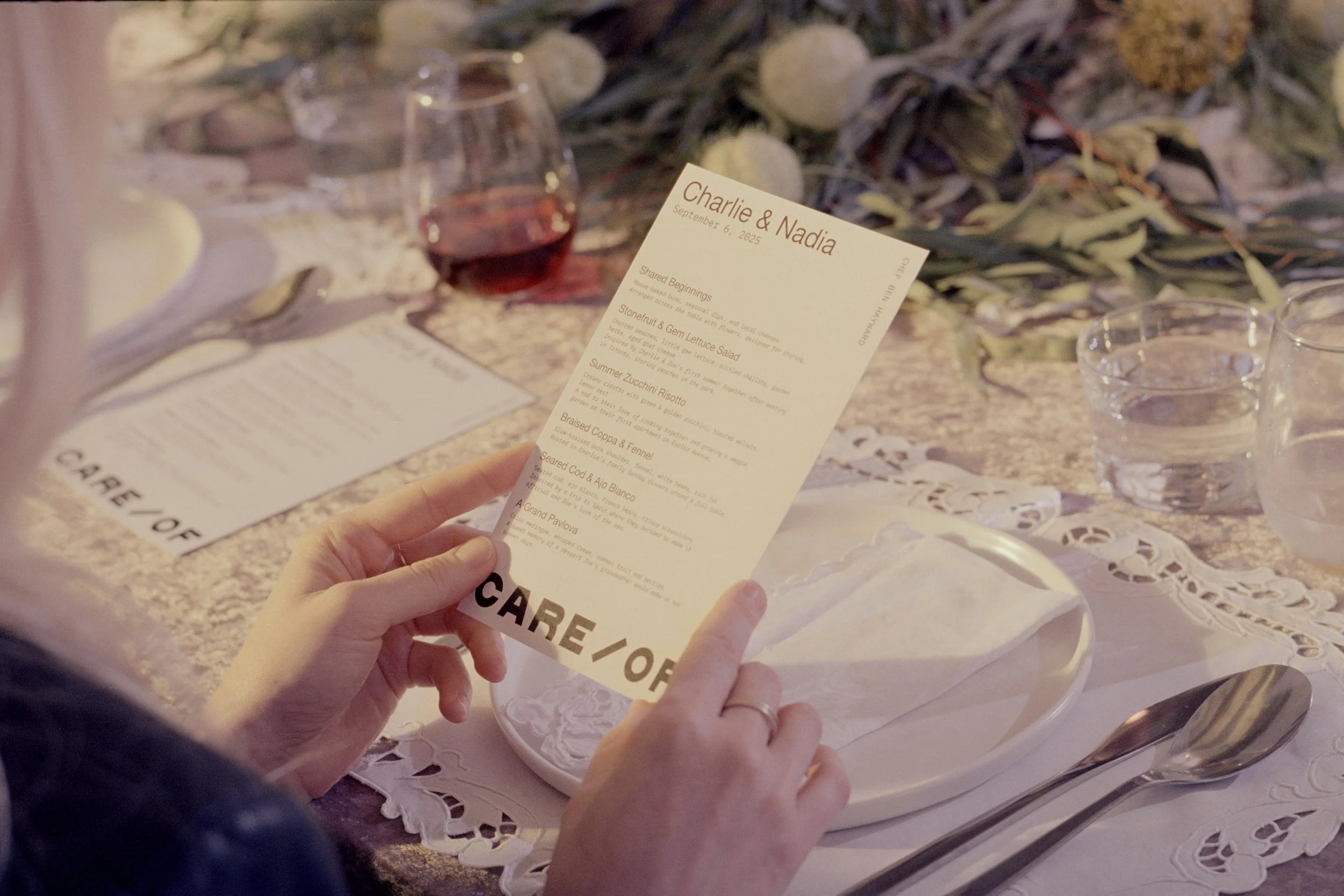 A person holding a printed menu at a dinner table set with plates, glasses of water, and wine. The table has a lace tablecloth and decorative greenery in the background.