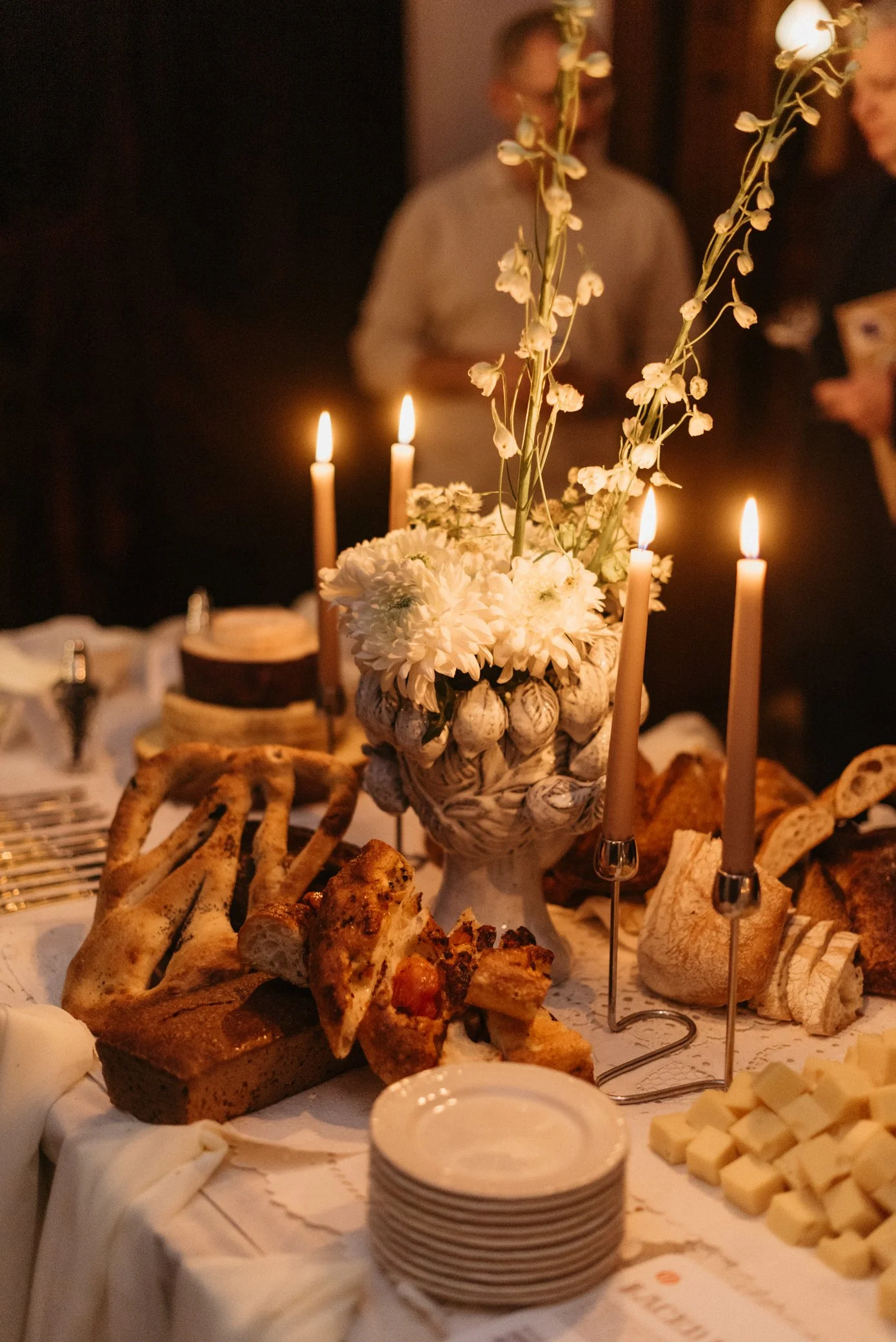 A table decorated with a floral centerpiece, lit candles, and a variety of bread and cheese at a celebration or gathering.
