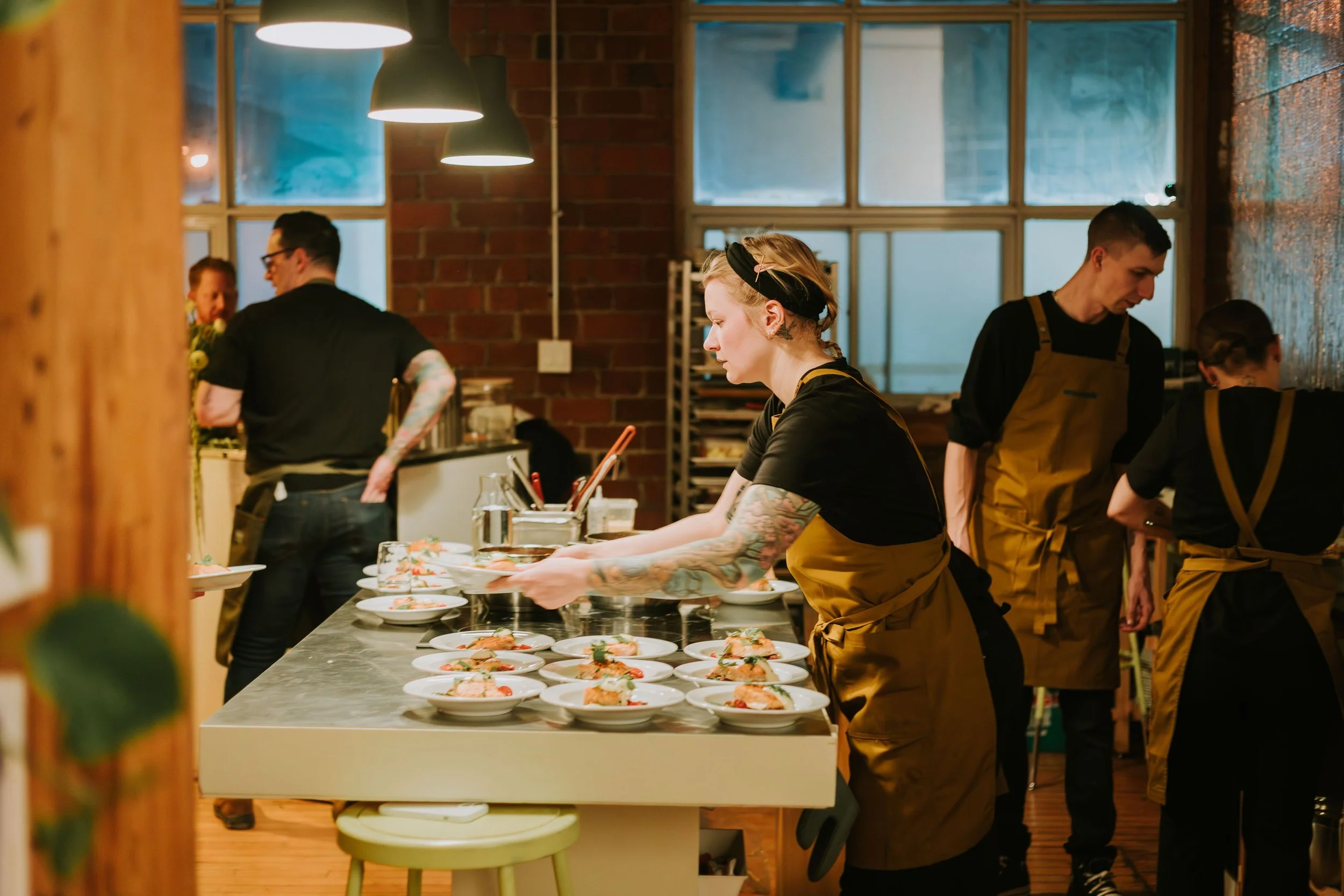 Chefs and kitchen staff preparing dishes in a restaurant kitchen with brick walls, large windows, and warm lighting.