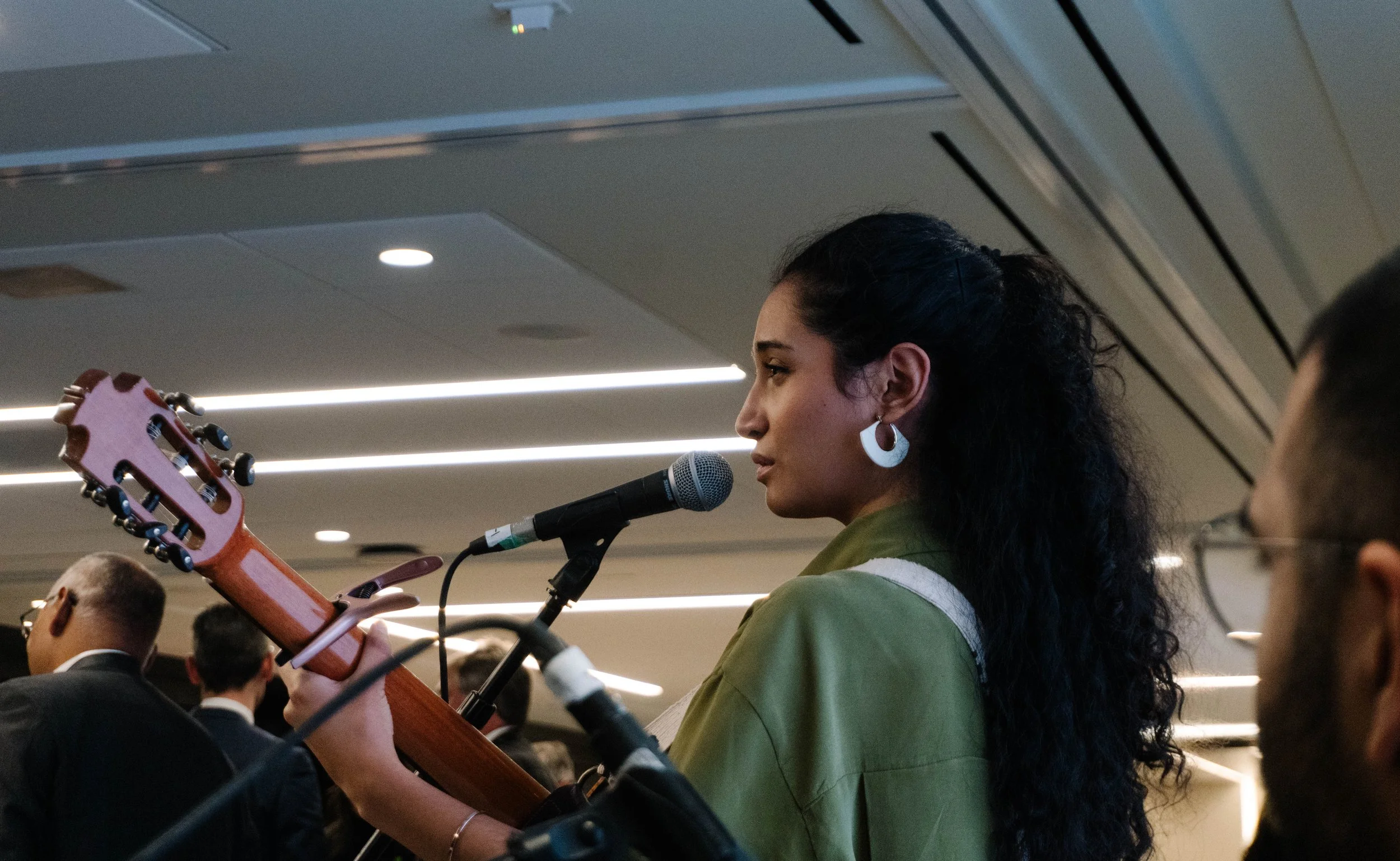 A woman with dark, curly hair is playing an acoustic guitar and singing into a microphone at an indoor event surrounded by other attendees.