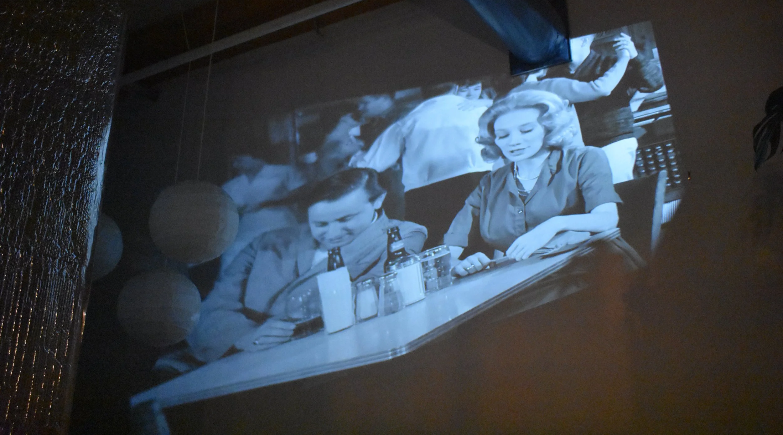 Black and white photo of two women sitting at a long table with drinks, smiling and looking down, in a busy environment with people in the background.