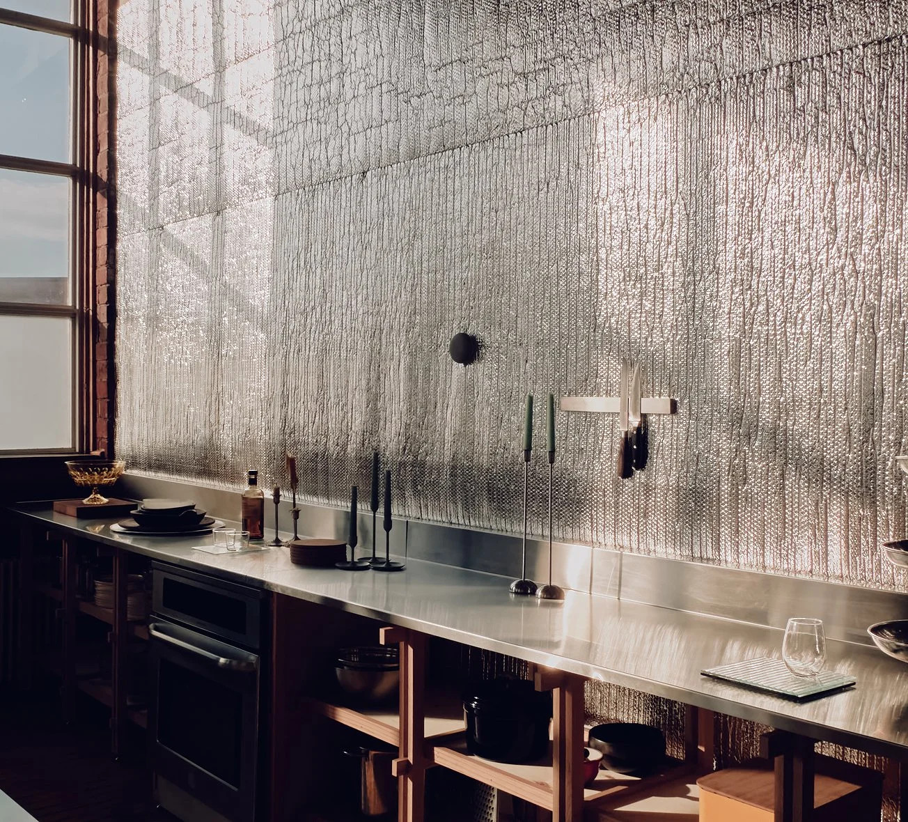 A kitchen with a long stainless steel countertop, various kitchen utensils, and dishes, with a textured metallic wall behind it and a window to the left.