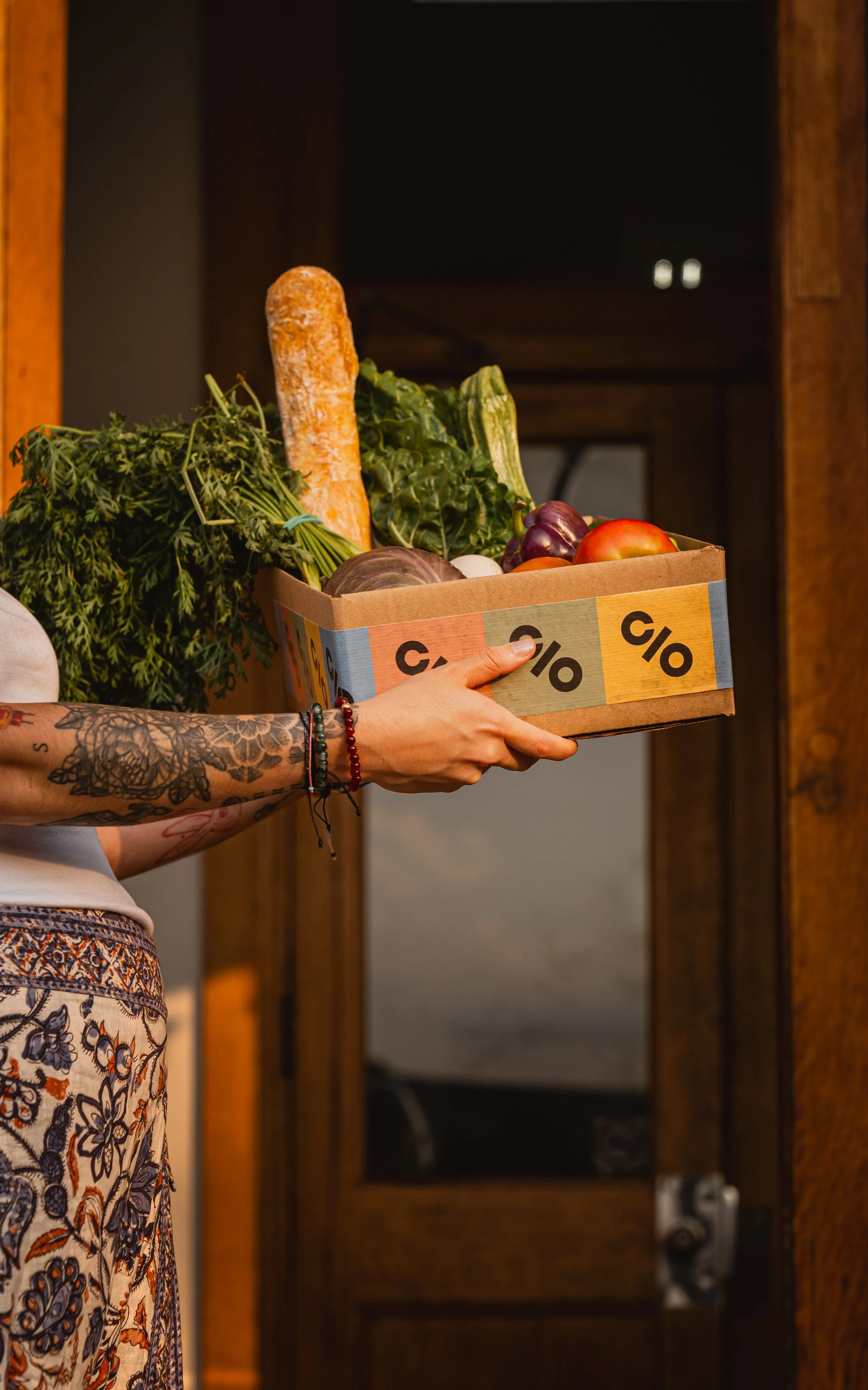 Person holding a cardboard box of fresh vegetables and bread, with a tattooed arm and jewelry, in front of wooden doors.
