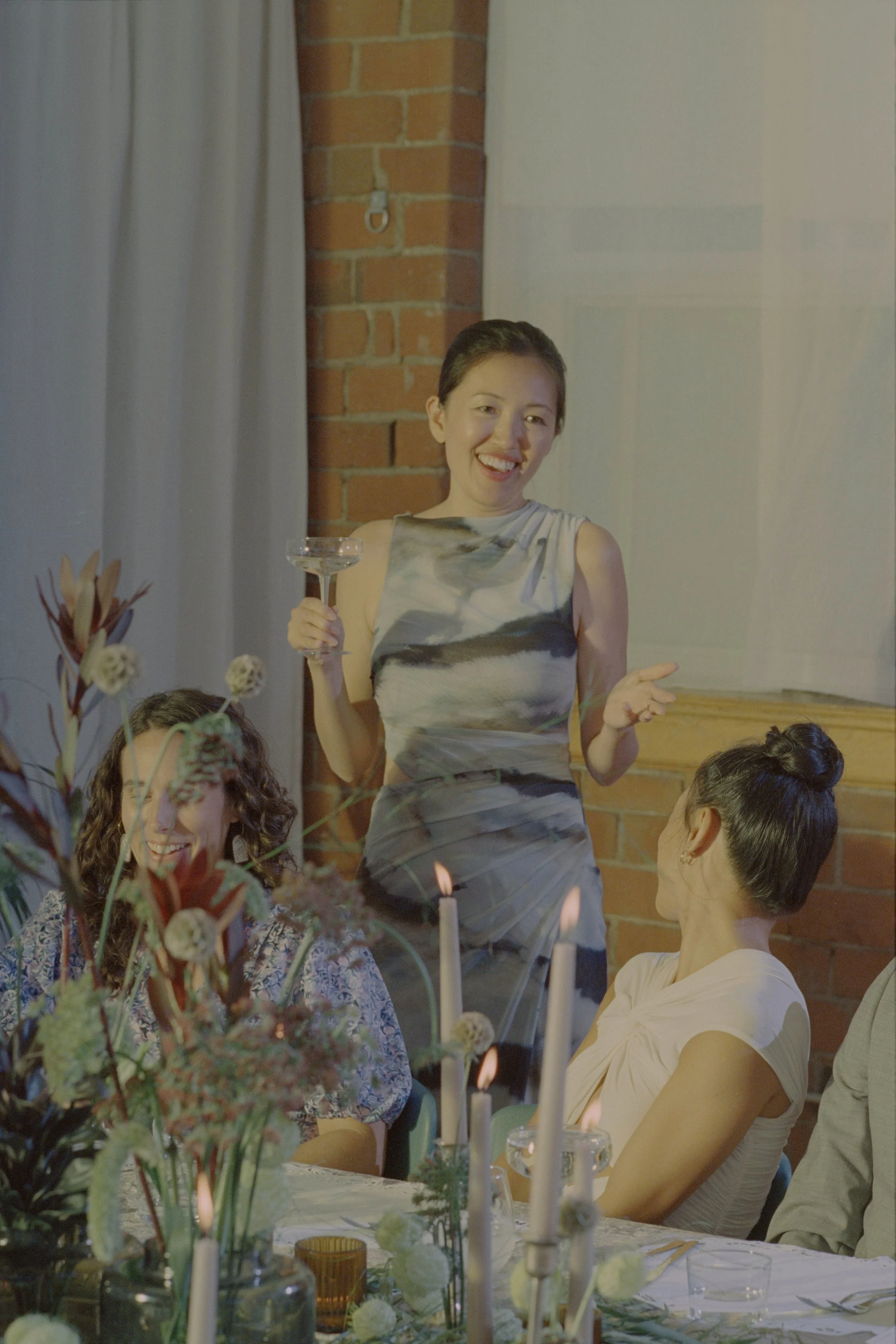 A woman in a sleeveless dress is standing, smiling, and holding a wine glass while speaking at a social gathering with other women seated at a table with floral arrangements and candles.