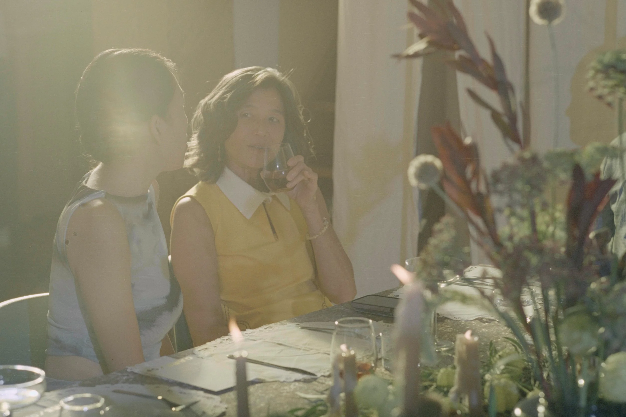 A woman in a yellow vest drinks red wine while sitting at a table with another woman during a gathering. The scene is softly lit with sunlight coming in from the side.