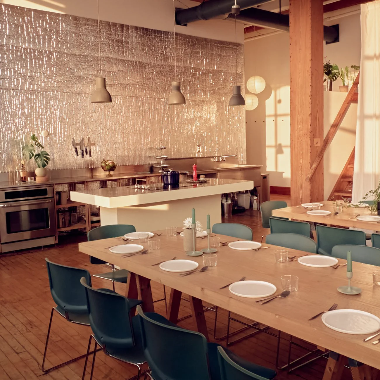A dining area with a long wooden table set for a meal, with plates, glasses, and candles, in front of a kitchen with hanging pendant lights, potted plants, and a shimmering textured wall in the background, with sunlight casting warm glows across the space.