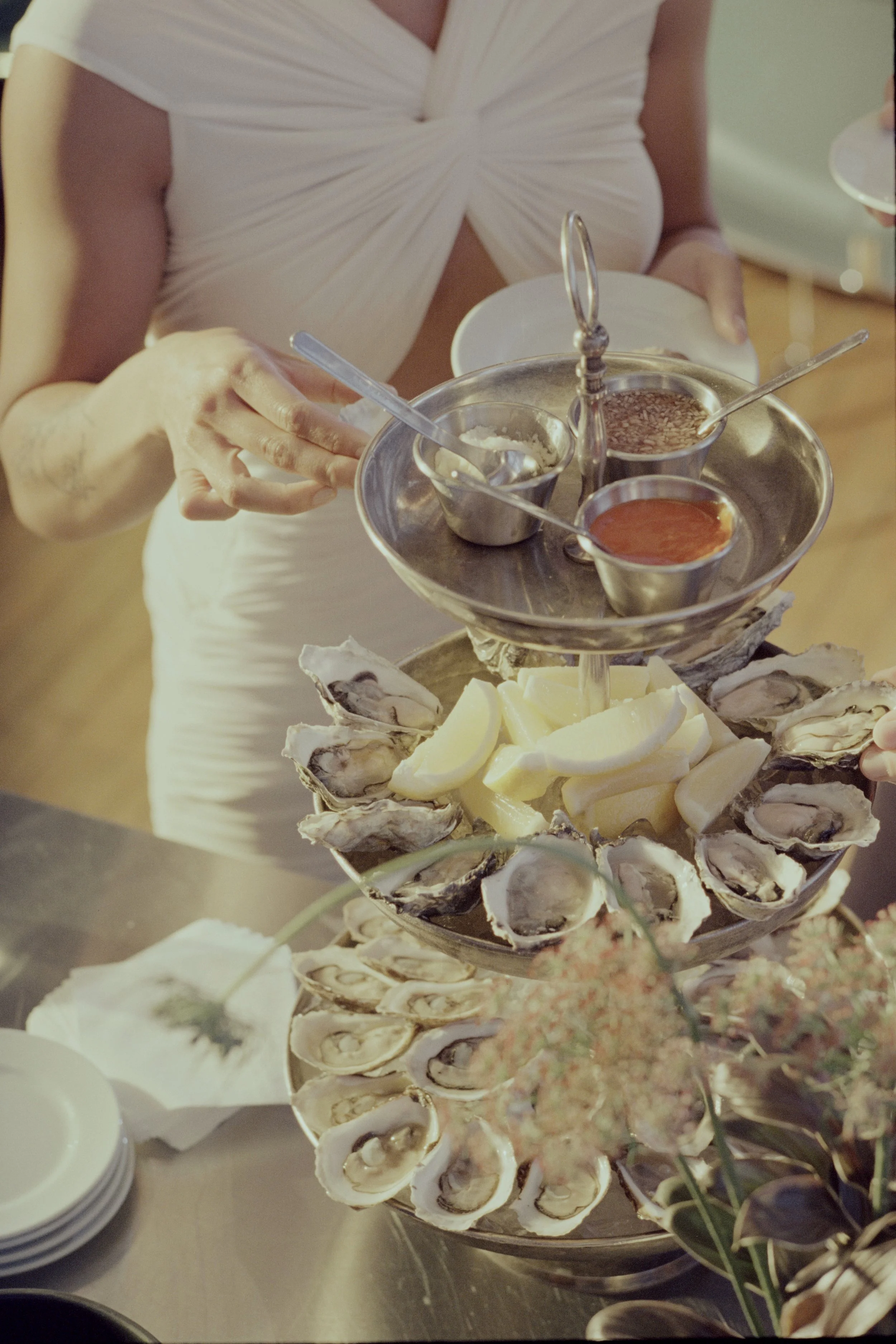 A tiered seafood platter with oysters, lemon wedges, and small bowls of sauces, topped with a woman in a white dress holding a plate.