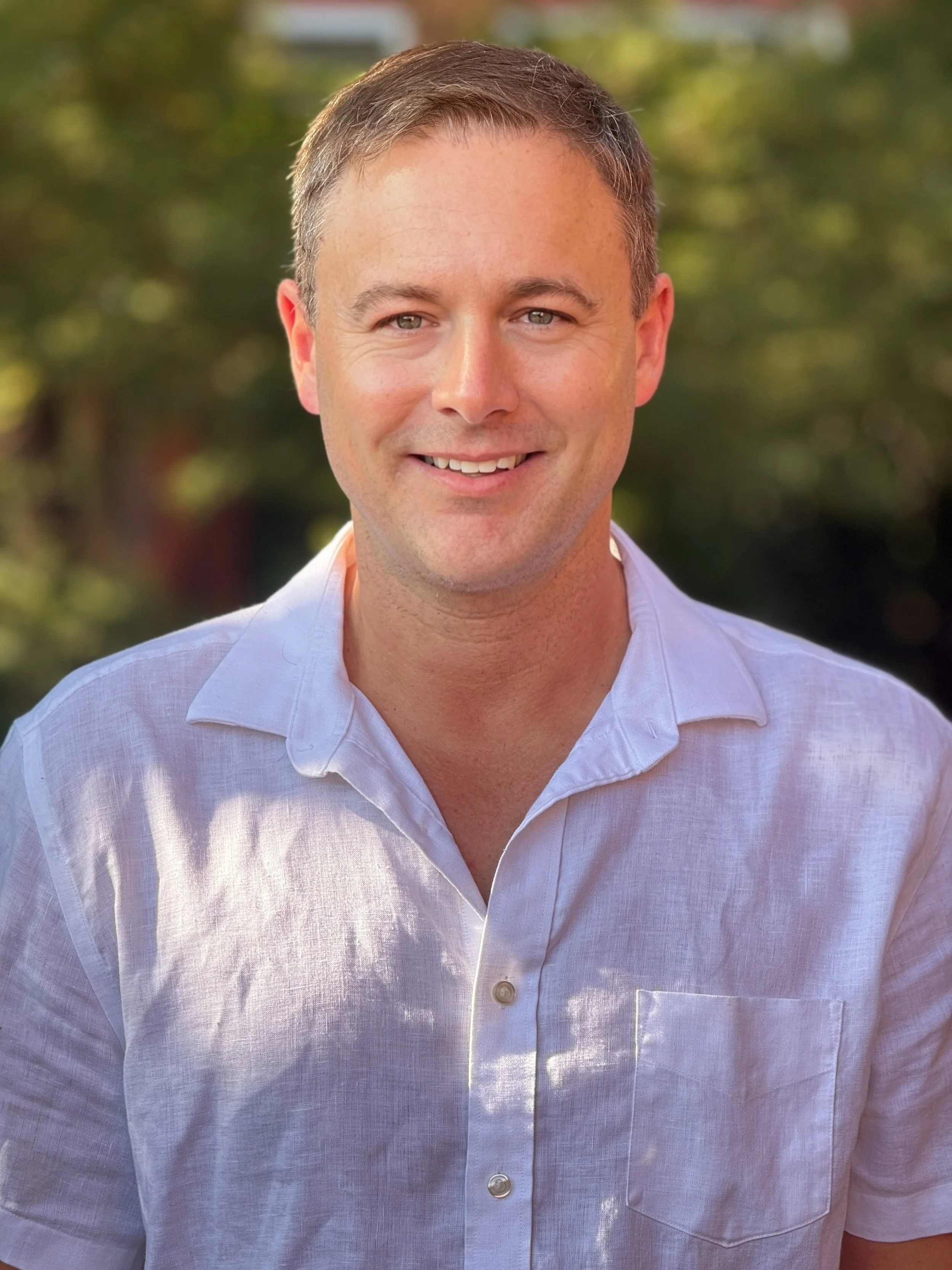 Portrait of a man with short light brown hair, light blue eyes, wearing a dark blue checkered shirt, standing outdoors with blurred green and autumn-colored foliage in the background.