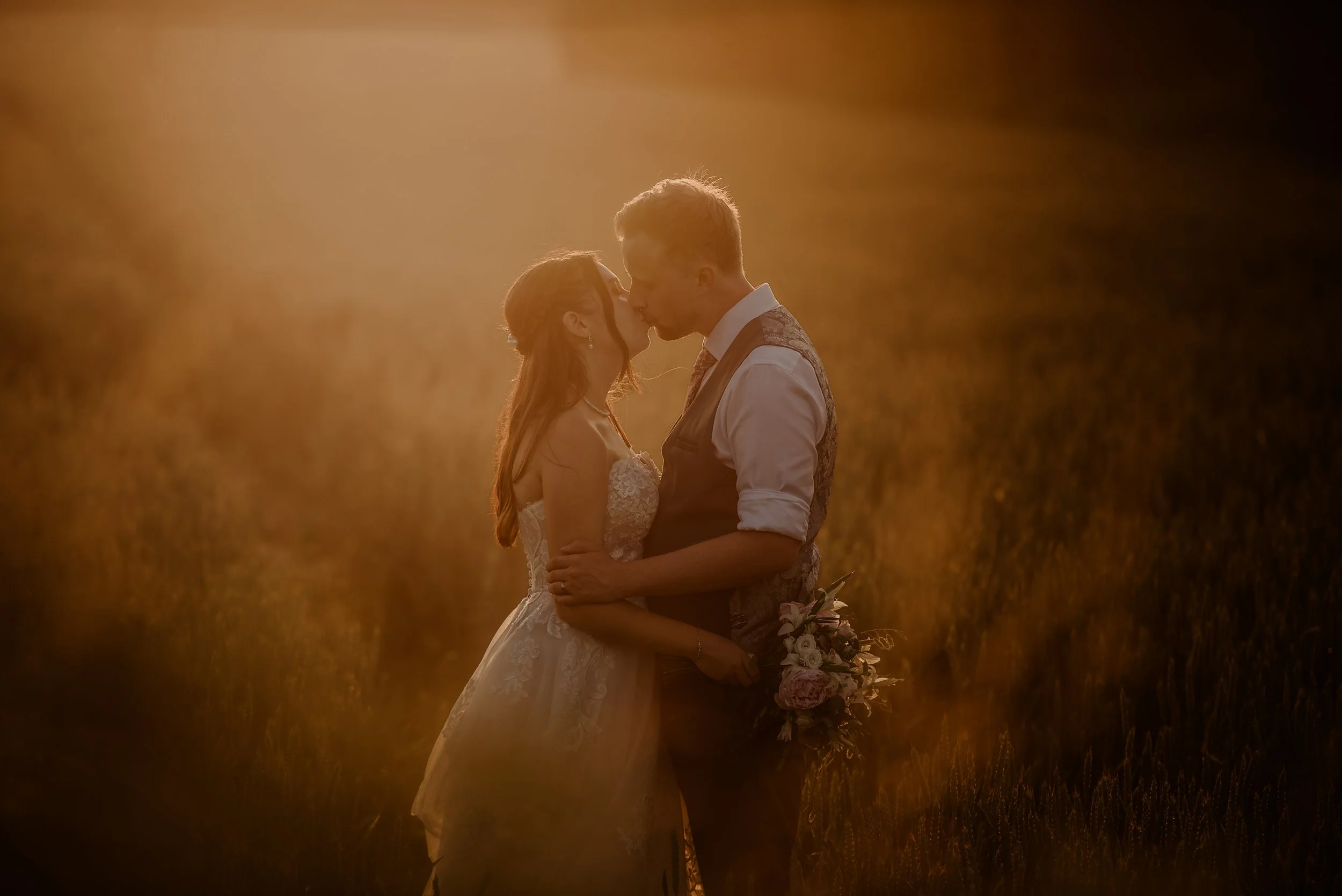 Bride and groom kissing in field at sunset opposite Old Kent Barn