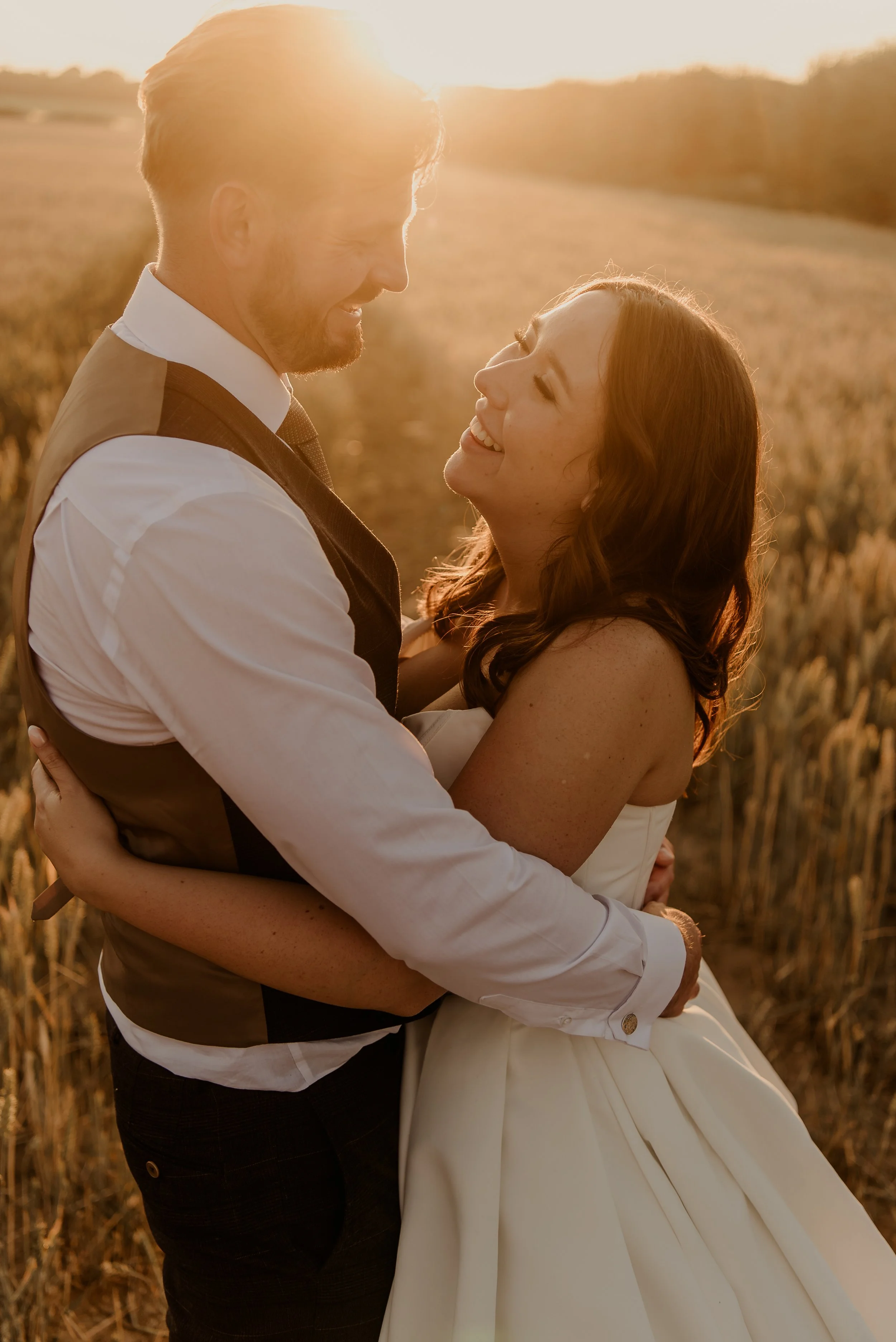 Bride and groom laughing whilst holding each other at sunset in a field opposite Old Kent Barn
