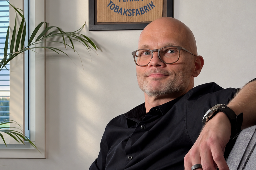 Rami Aaltonen istuu sivuttain harmaalla sohvalla mustassa kauluspaidassa. Ikkunasta tulee luonnonvaloa.