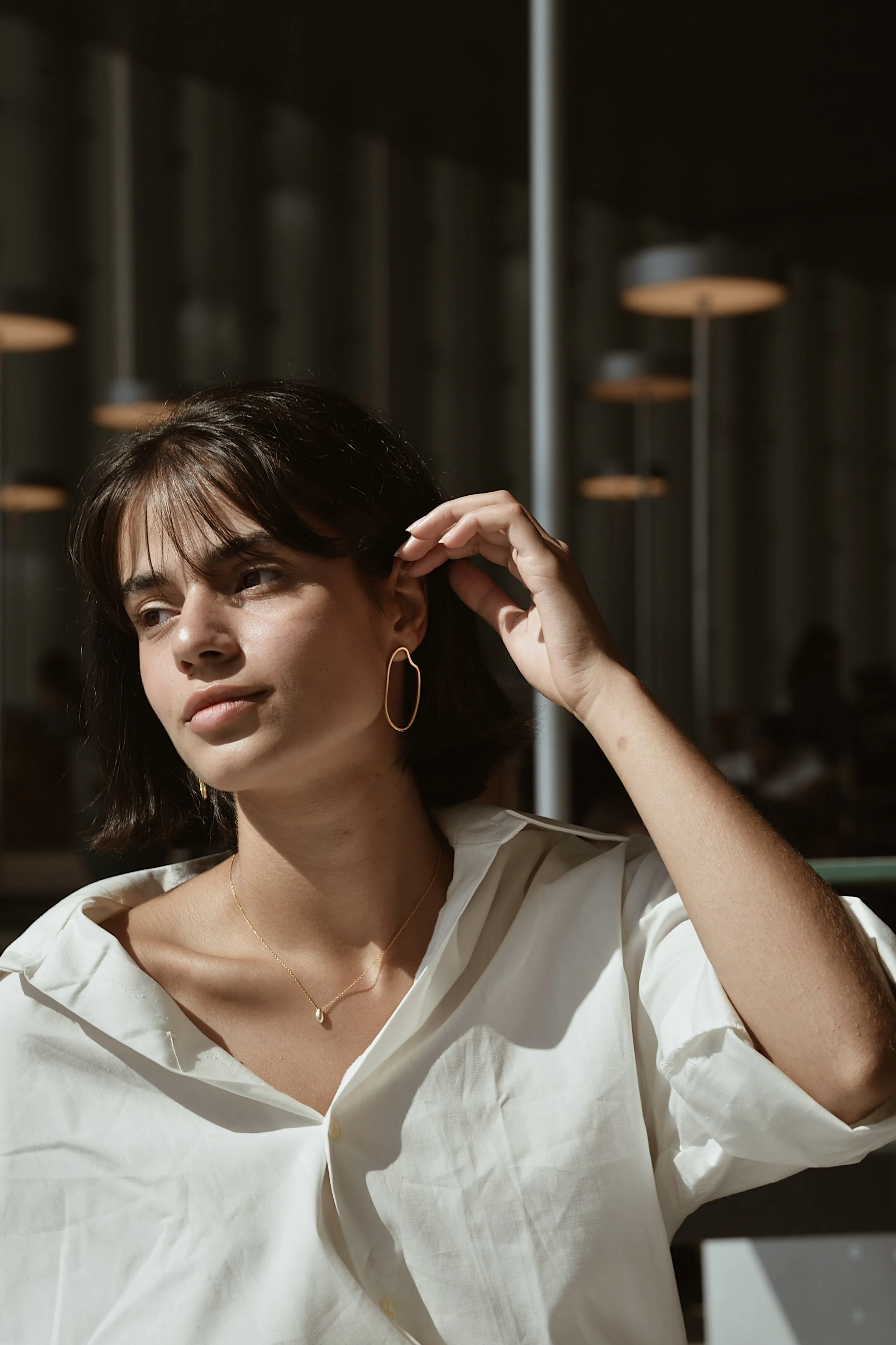 A young woman with short dark hair wearing a white blouse, gold hoop earrings, and a delicate gold necklace, sitting indoors at a table in the sunlight.