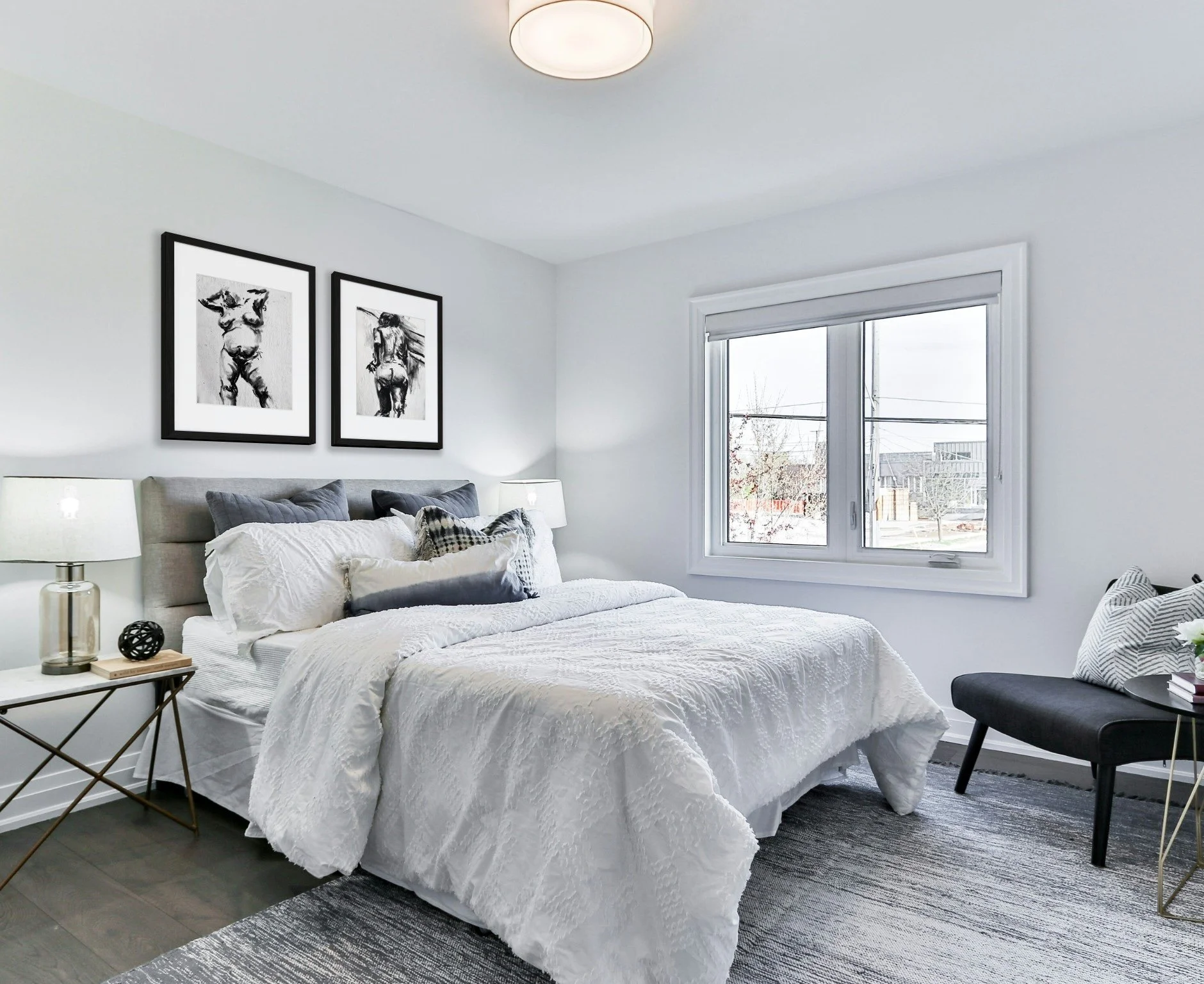 A modern bedroom with a neatly made white bed, gray pillows, and black-and-white artwork of a dog and a person on the wall behind the bed. There are two white lamps, a black bench with a pillow and a plant on a side table near the window. The room has a large window showing an outdoor scene with trees and a building.
