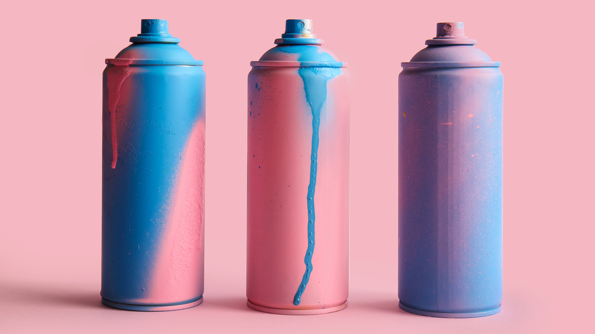 Three spray paint cans with pink and blue paint against a pink background.