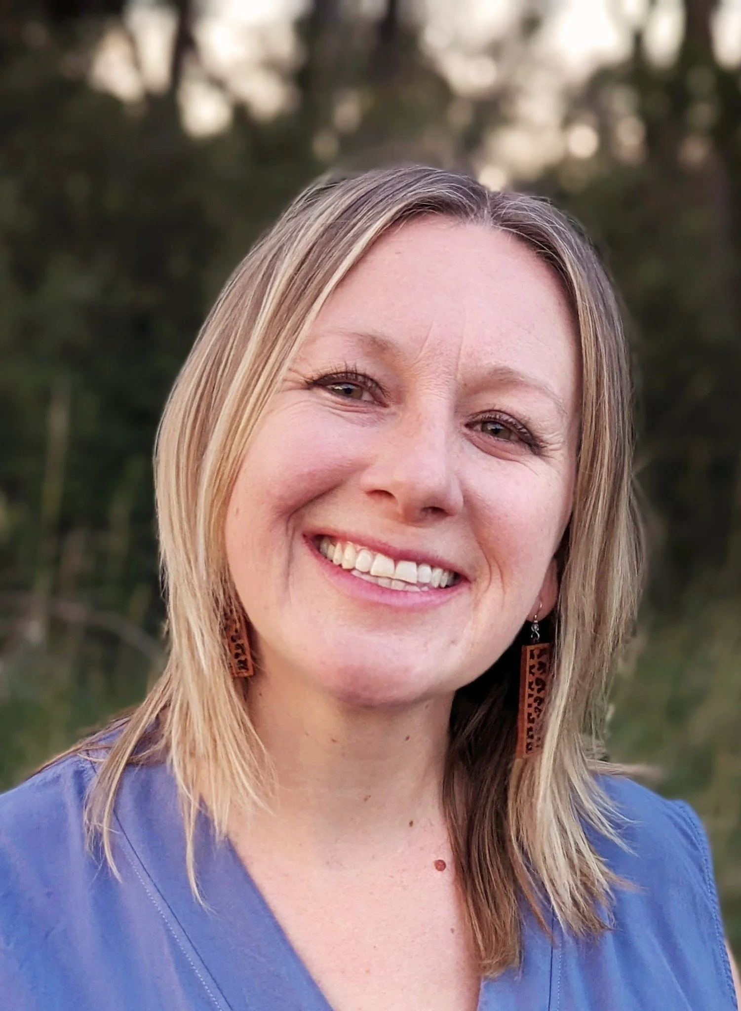 A woman with blonde hair smiling outdoors, wearing a blue shirt and brown earrings, with blurred green trees in the background.