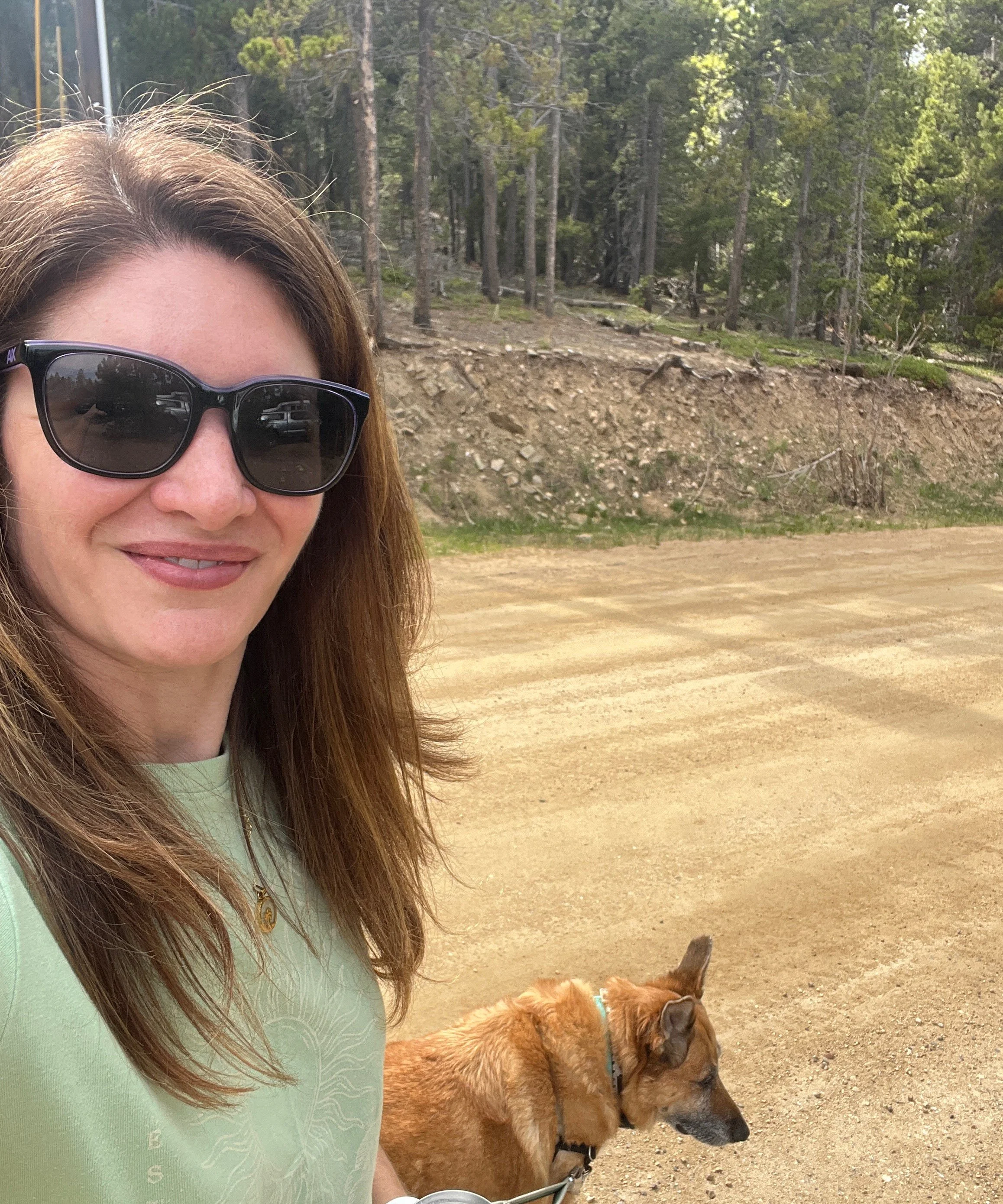 A woman with long brown hair wearing black sunglasses and a light green shirt, standing outdoors on a dirt path with a brown dog on a leash, in a forested area with trees in the background.