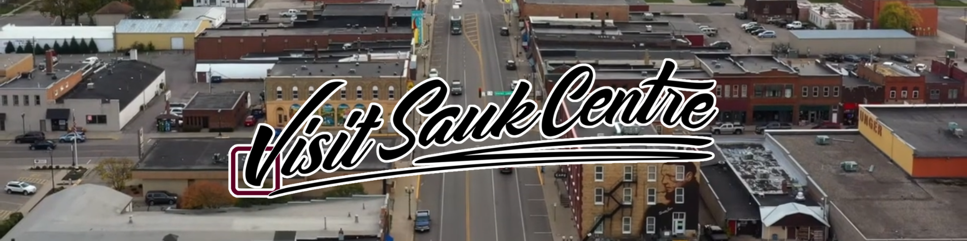 Aerial view of downtown Sauk Centre showing streets, buildings, parking lots, and cars with the text "Visit Sauk Centre" overlaid.