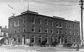 Black and white photo of a three-story brick building on a street corner, with multiple windows and an entrance at street level. The building has a flat roof and is situated in an urban setting with power lines and streetcars visible.