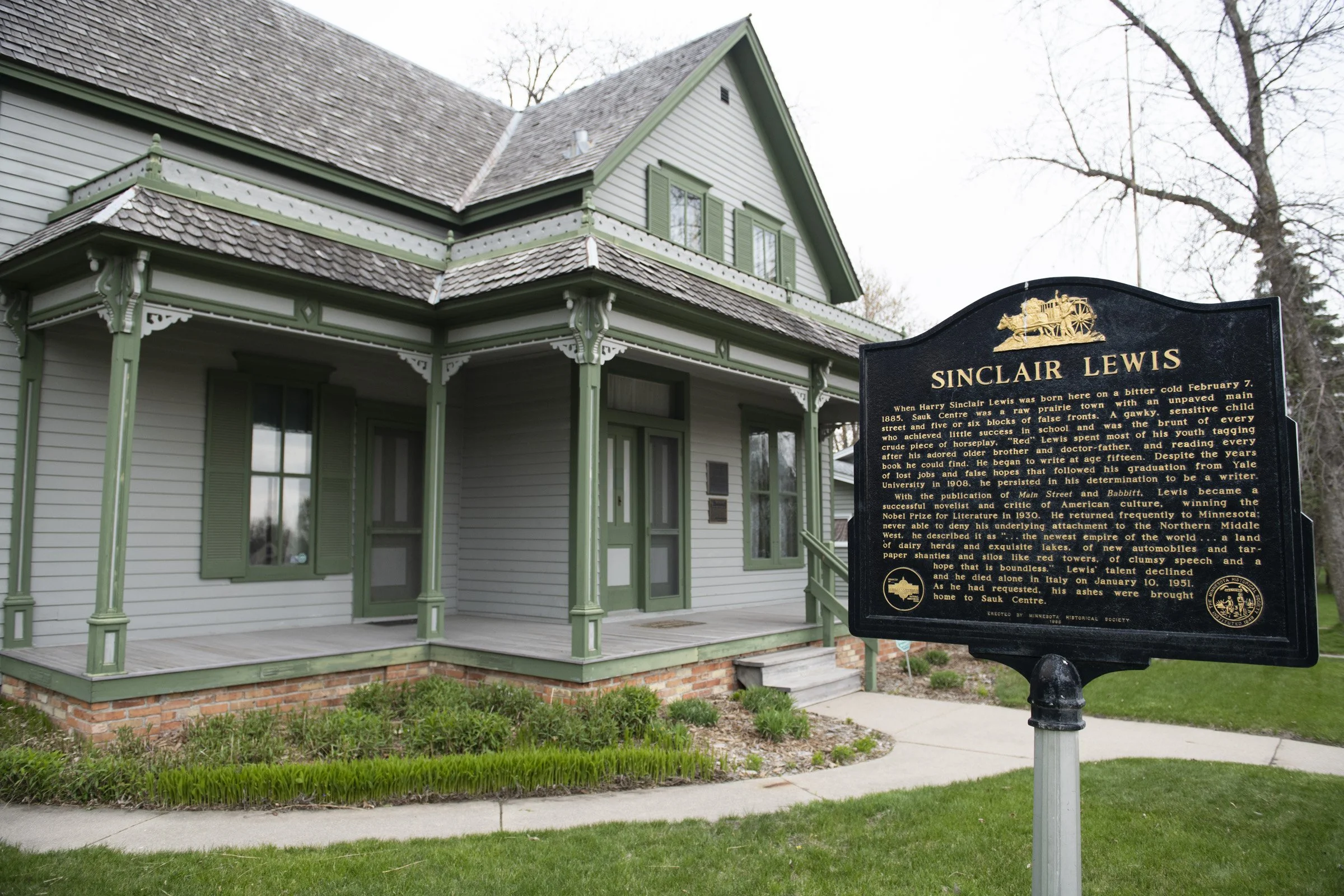 Historic house with green trim and a porch, located at Sioux Center. A black historical marker in front describes Sinclair Lewis, an American novelist born in the area.