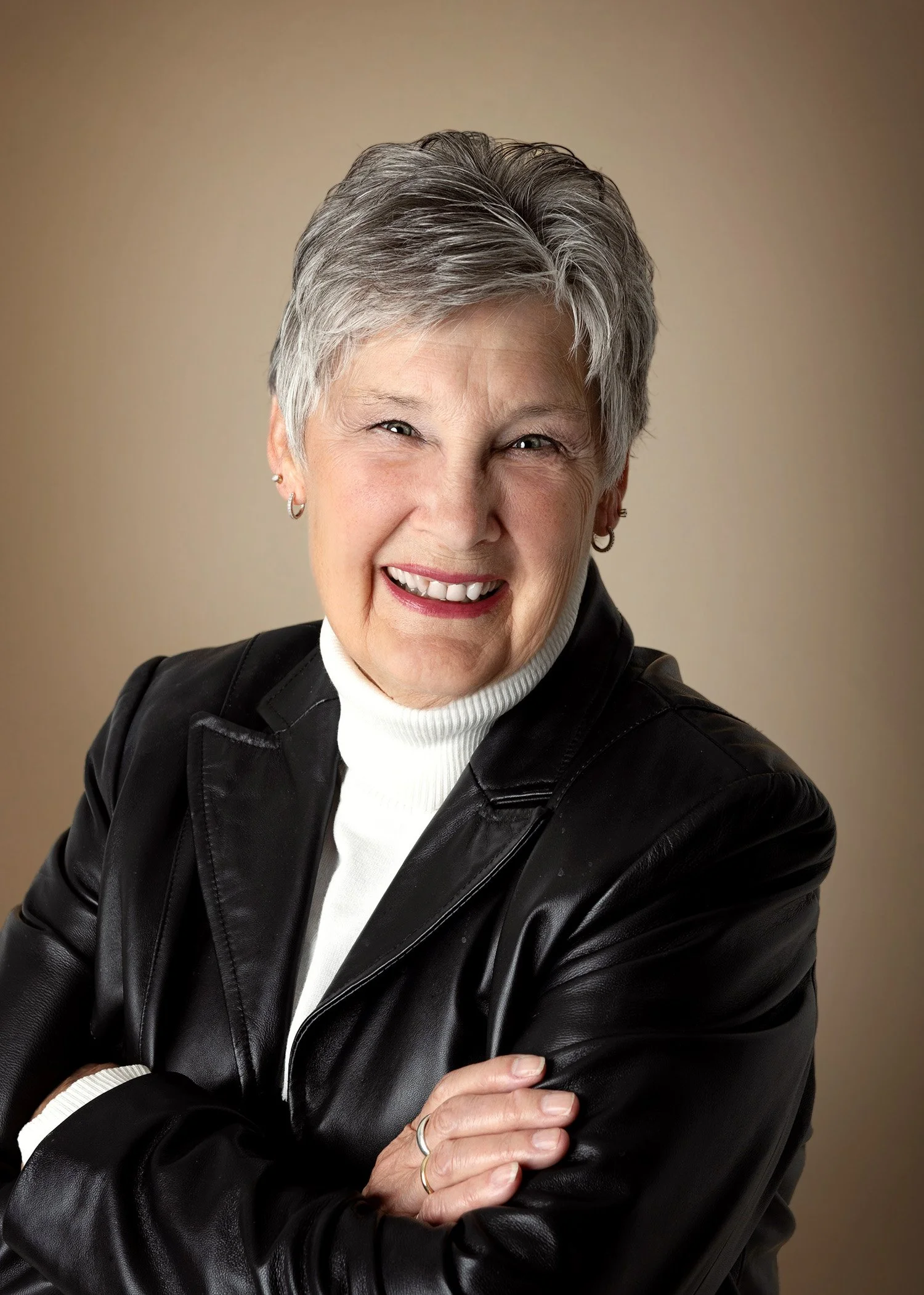 Portrait of an older woman with short gray hair, smiling, wearing a black leather jacket over a white turtleneck, with her arms crossed.