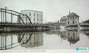 Historical black-and-white photo of a riverfront with industrial buildings and a small pavilion, reflecting in the water.